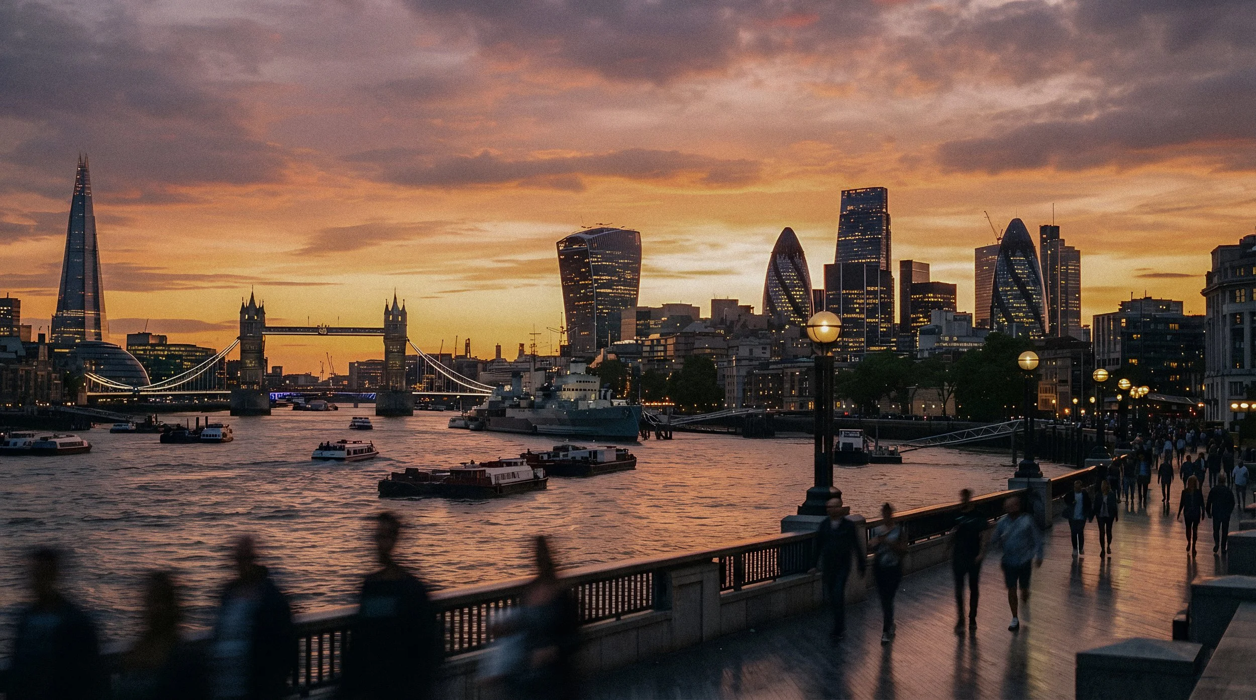 Sunset over the London skyline with iconic buildings including The Shard, City Hall, and the Tower Bridge, as seen from a riverside promenade with people walking and boats on the River Thames.