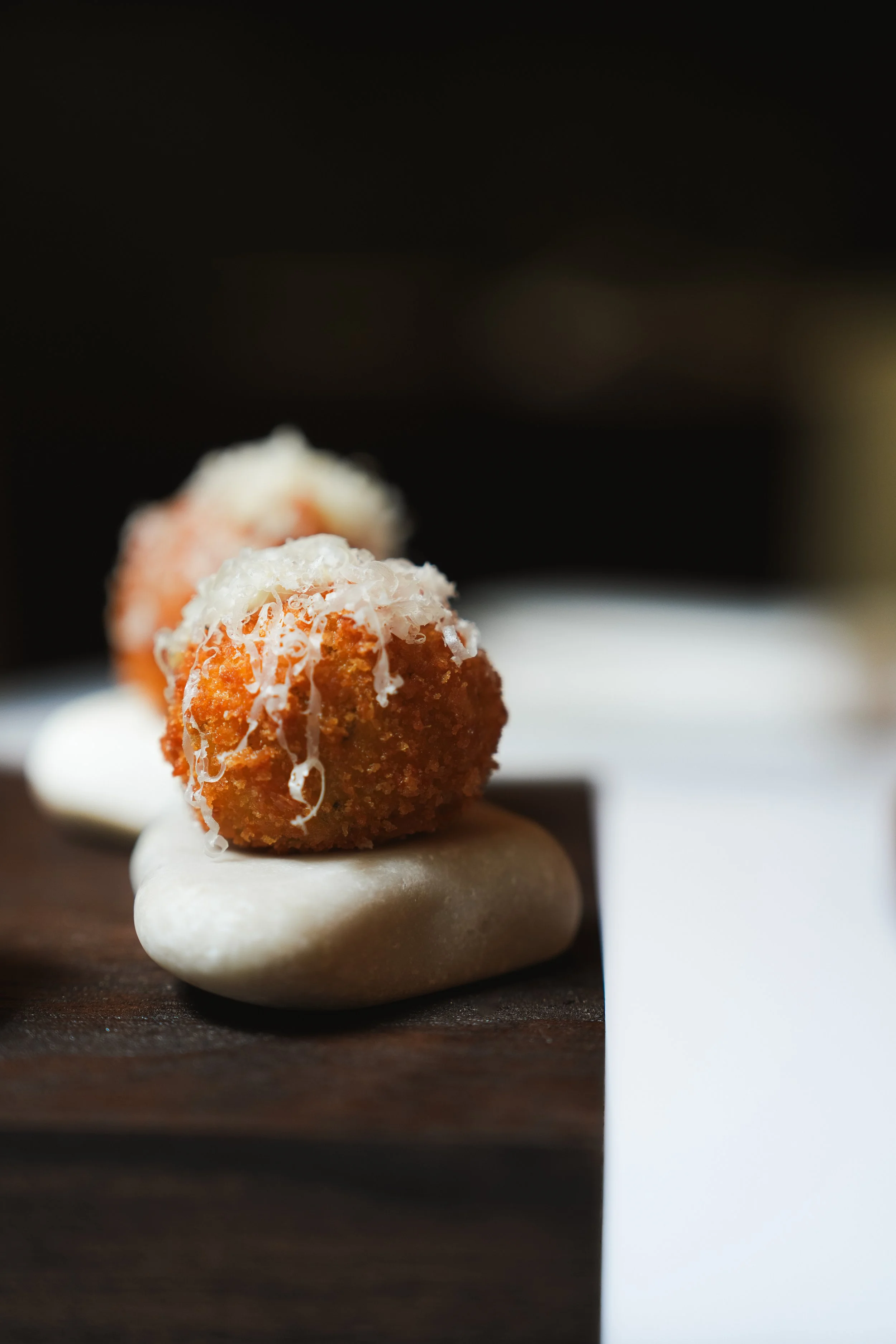 Close-up of two breaded fried balls topped with grated cheese on a plate, with a dark background.