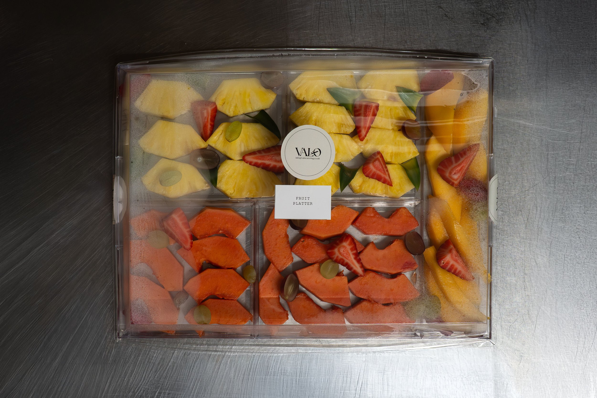 A clear plastic fruit platter containing cut pineapples, strawberries, grapes, watermelon, cantaloupe, and yellow bell pepper slices, with a label that reads 'VALO' and 'fruit platter'.