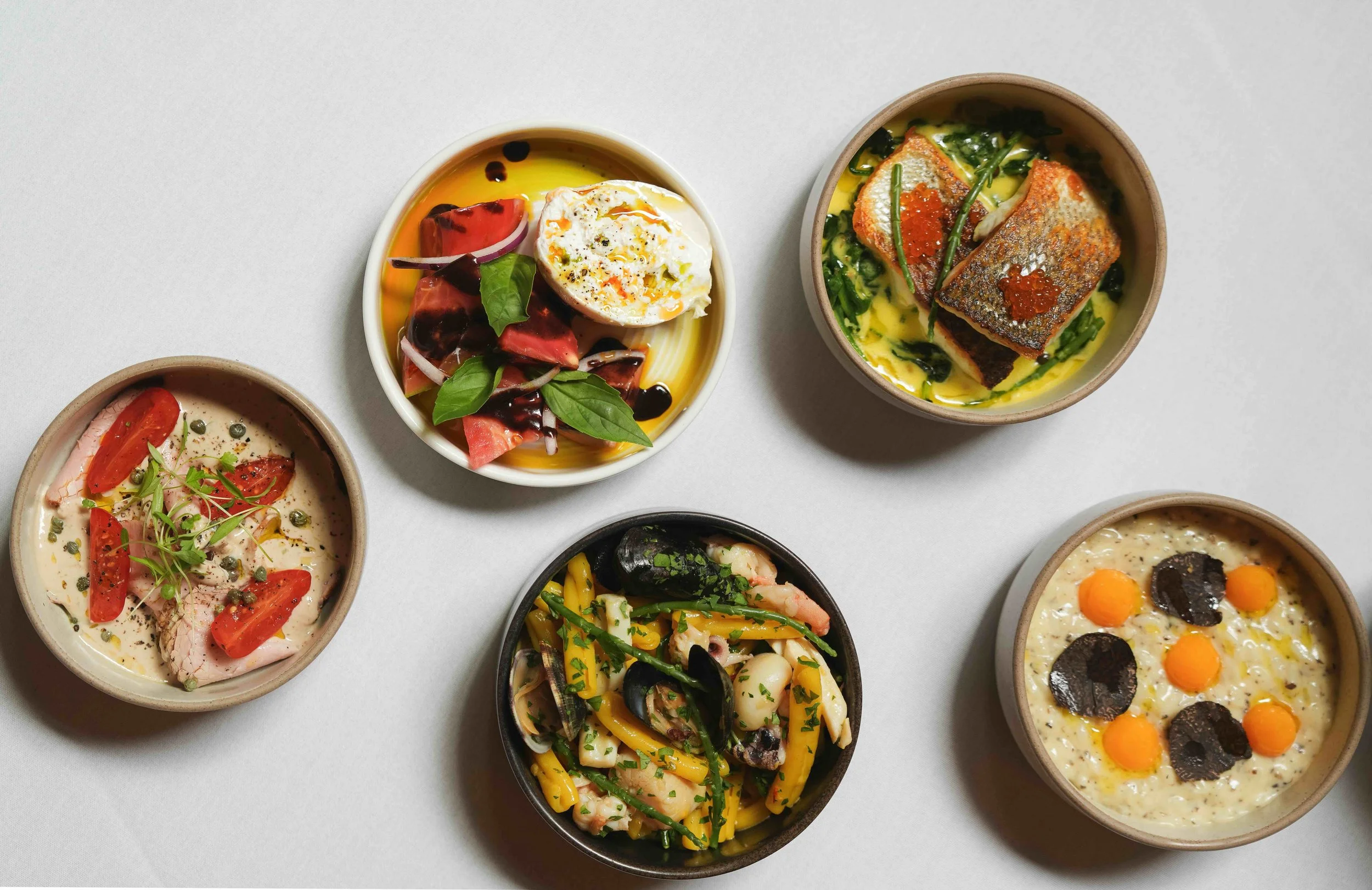 Assorted small bowls of colorful dishes including salads, eggs, vegetables, and creamy spreads on a white surface.
