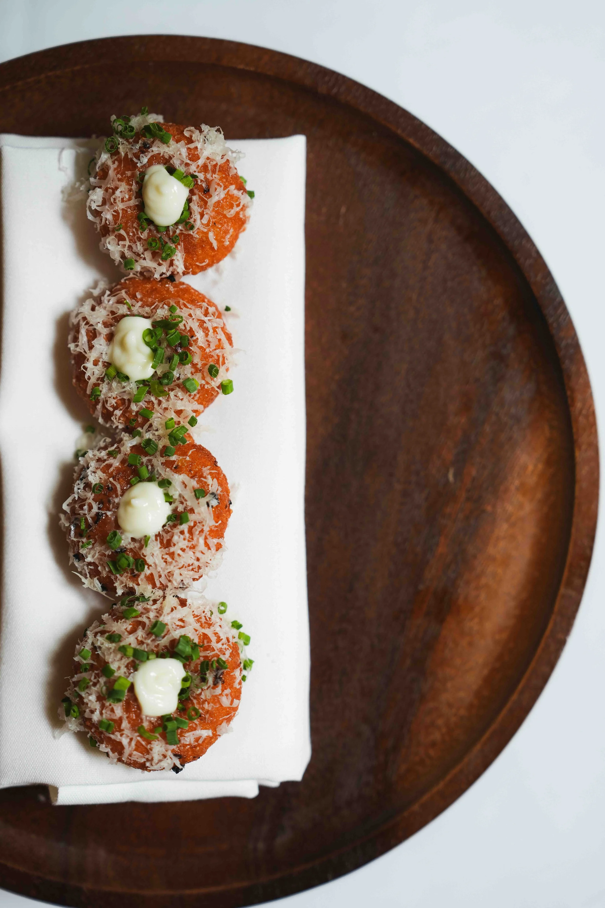 Three fried appetizers coated in orange sauce, topped with grated cheese, small dollops of white sauce, and chopped green onions, served on a white plate with a wooden tray underneath.