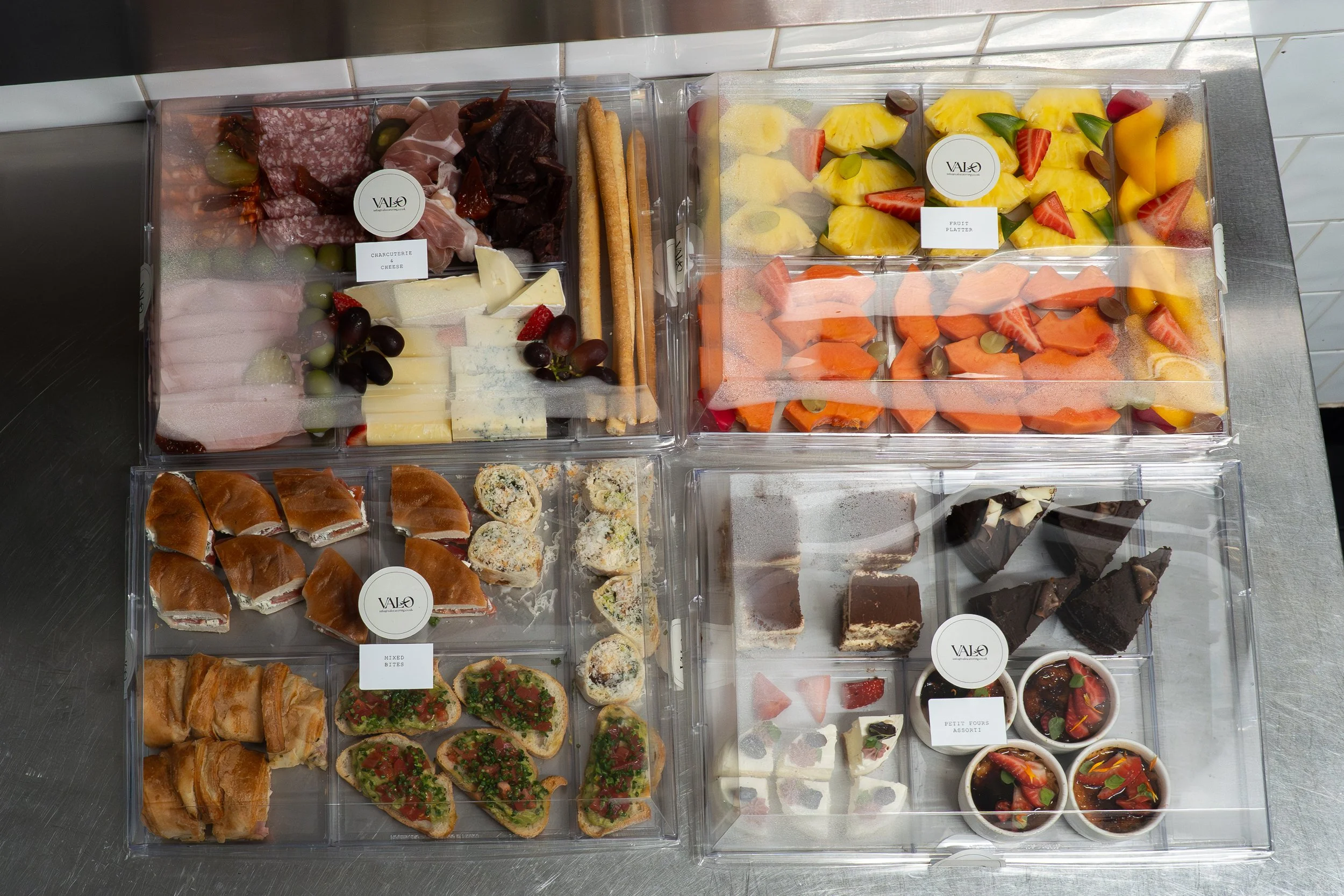 Assorted food trays with charcuterie, cheese, bread, and fruit, including strawberries, pineapple, and melon, labeled with small tags, displayed in clear containers.