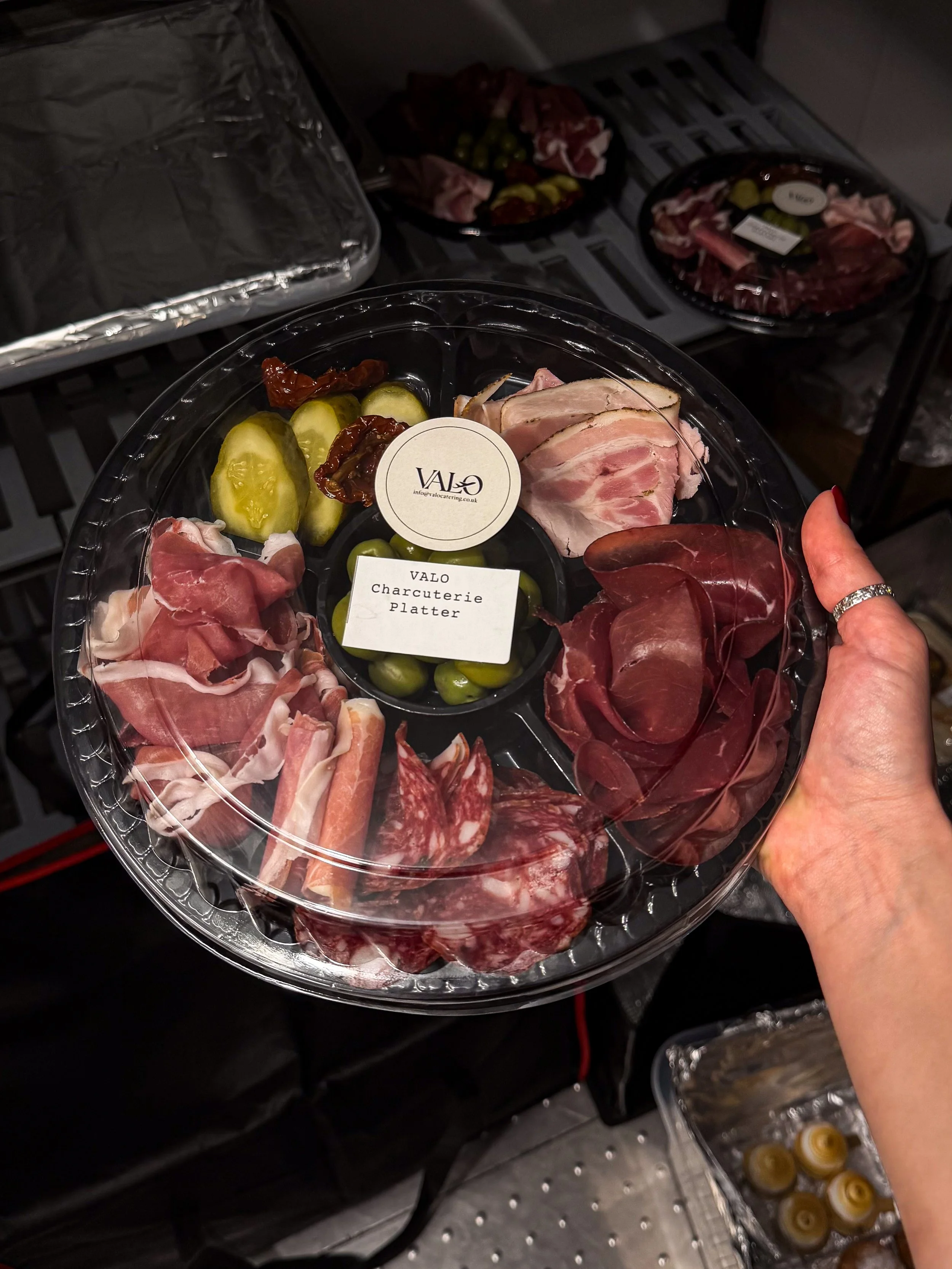 Person holding a plastic tray with an assortment of sliced meats, pickles, and sun-dried tomatoes, labeled 'VALO Charcuterie Platter,' in a kitchen setting.