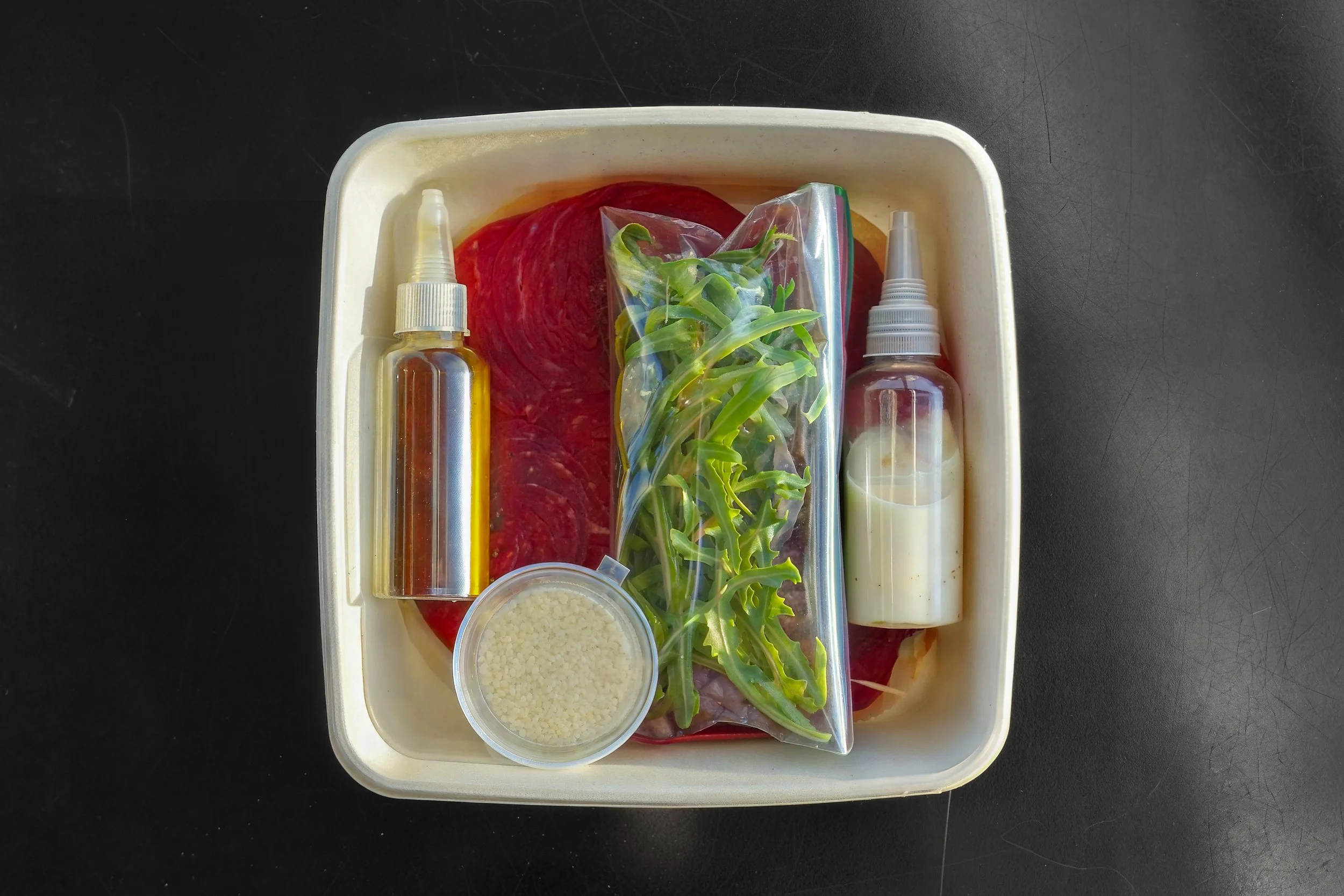 A plastic container holding various ingredients including raw meat, green leafy herbs, a container of grated cheese, and two squeeze bottles with liquids, set against a black background.