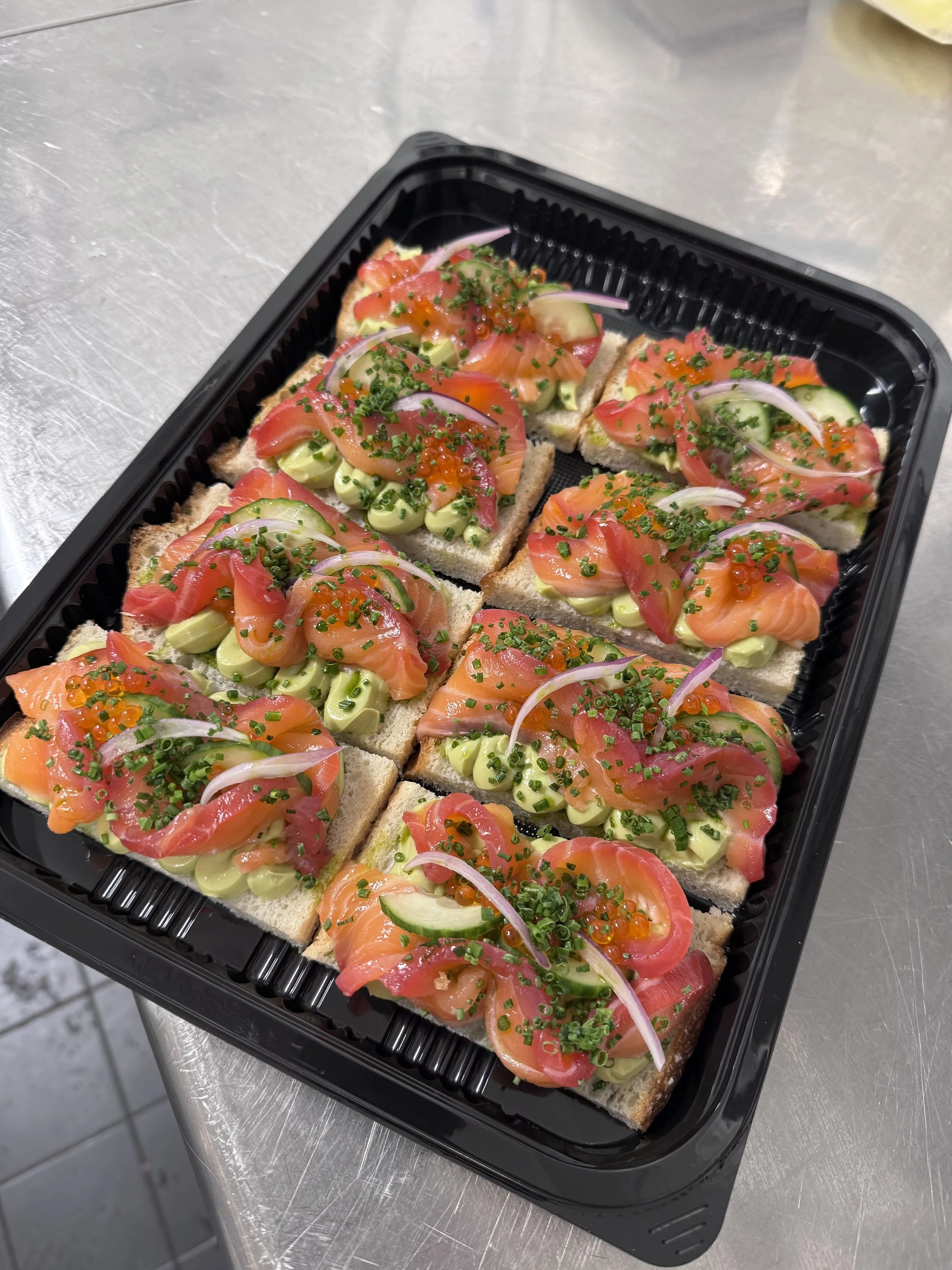 Platter of handcrafted smoked salmon and avocado open sandwiches topped with cucumber, red onion, fresh chives, and salmon roe, freshly prepared for private jet catering.