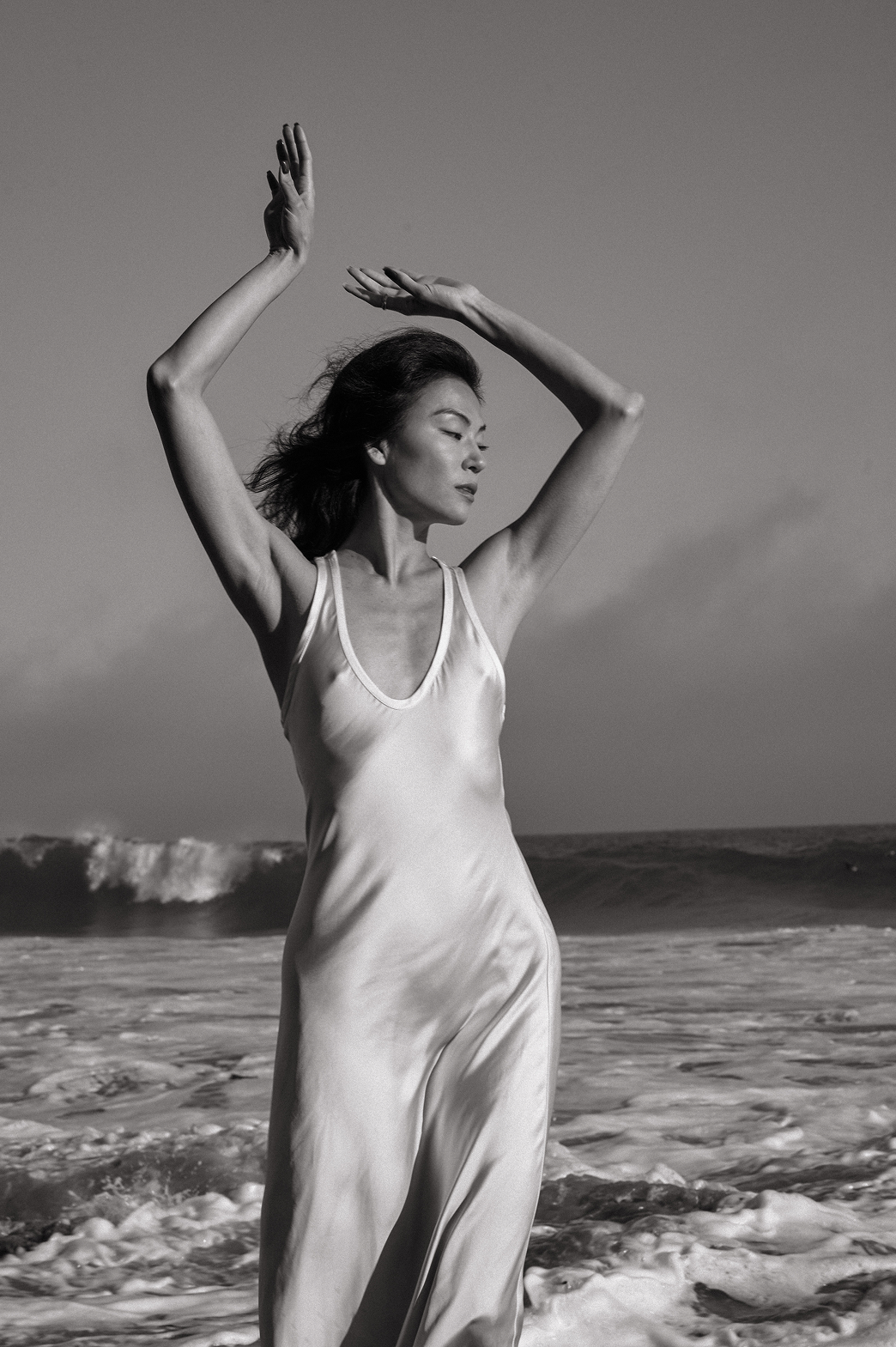 A woman in a light-colored dress standing on the beach with waves in the background, her arms raised above her head, eyes closed, and hair flowing in the wind, in black and white photography.