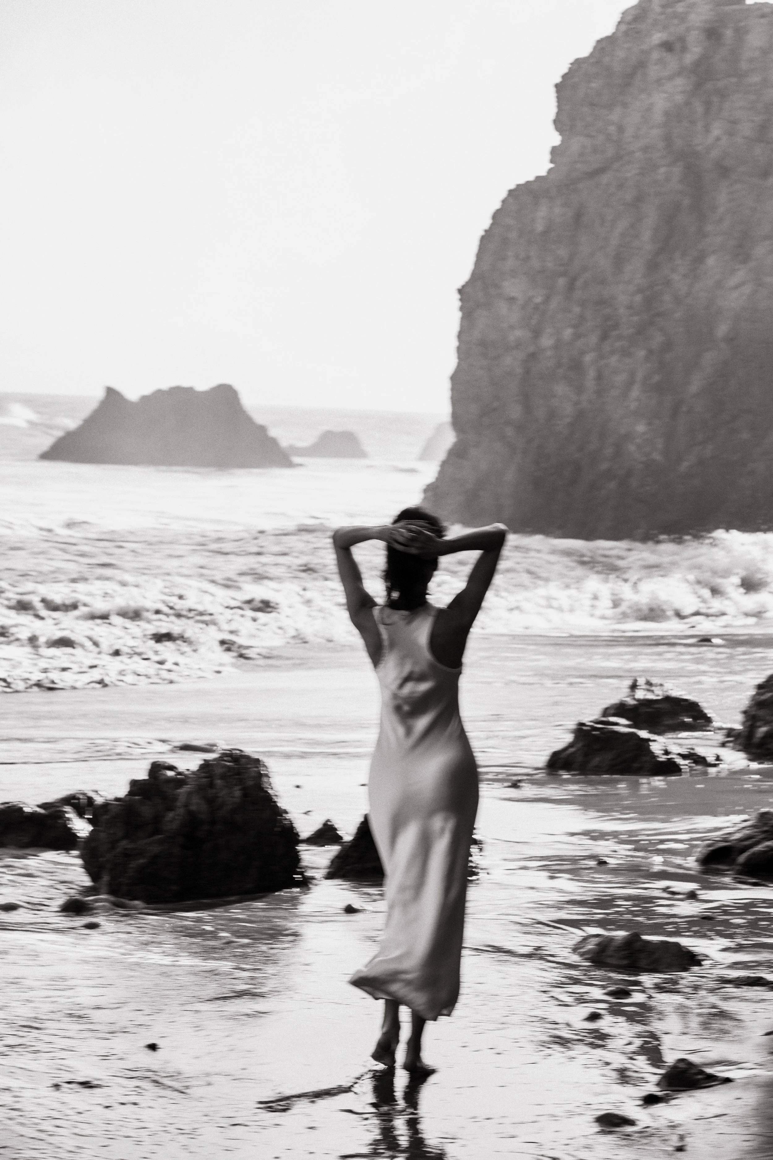 A woman in a long dress stands in the shallow water of a beach, with her hands behind her head, facing the ocean. Large rocks and cliffs are visible in the background, with a hazy sky overhead.
