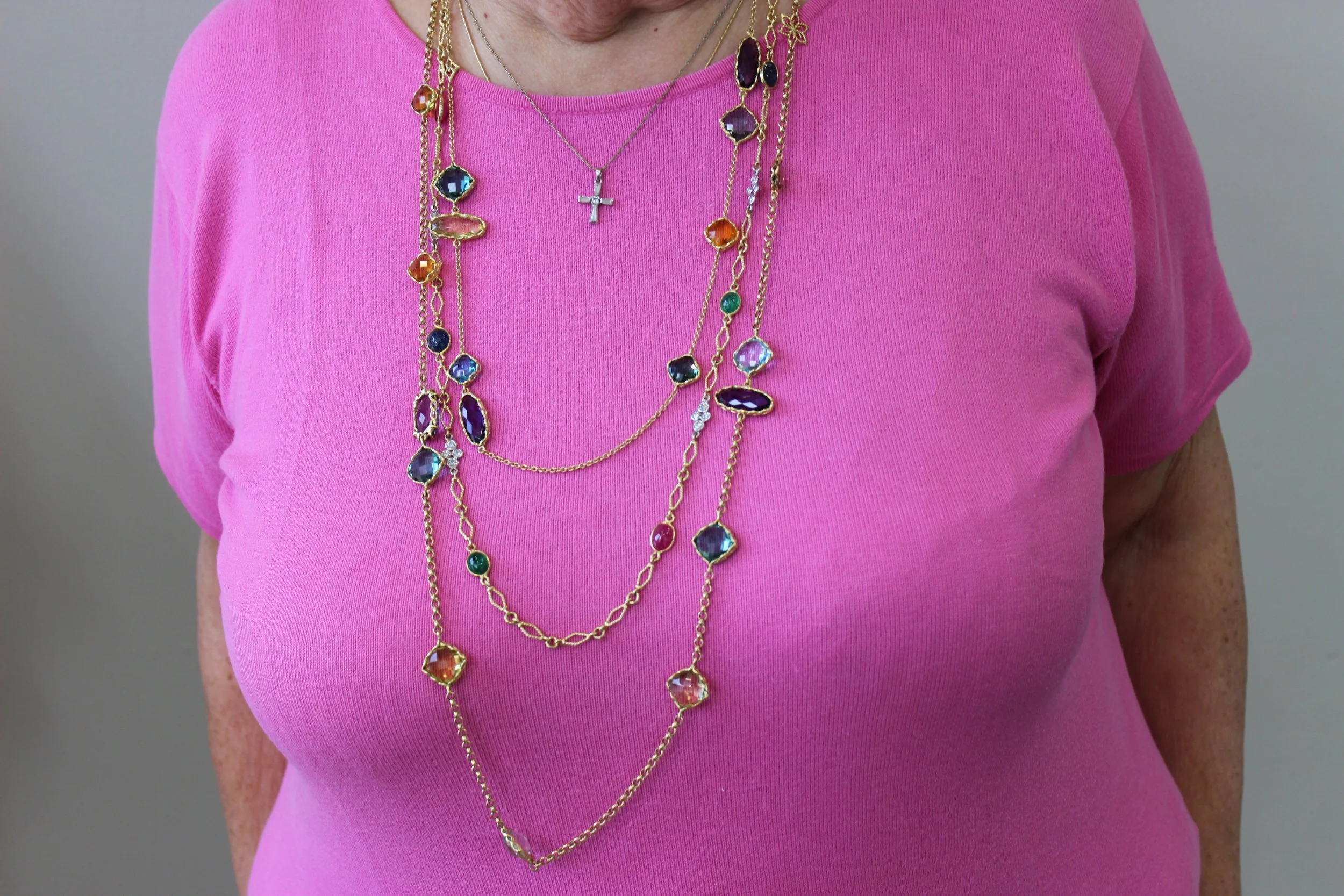 A woman wearing a pink top with multiple layered gold necklaces adorned with colorful gemstones and a cross pendant.