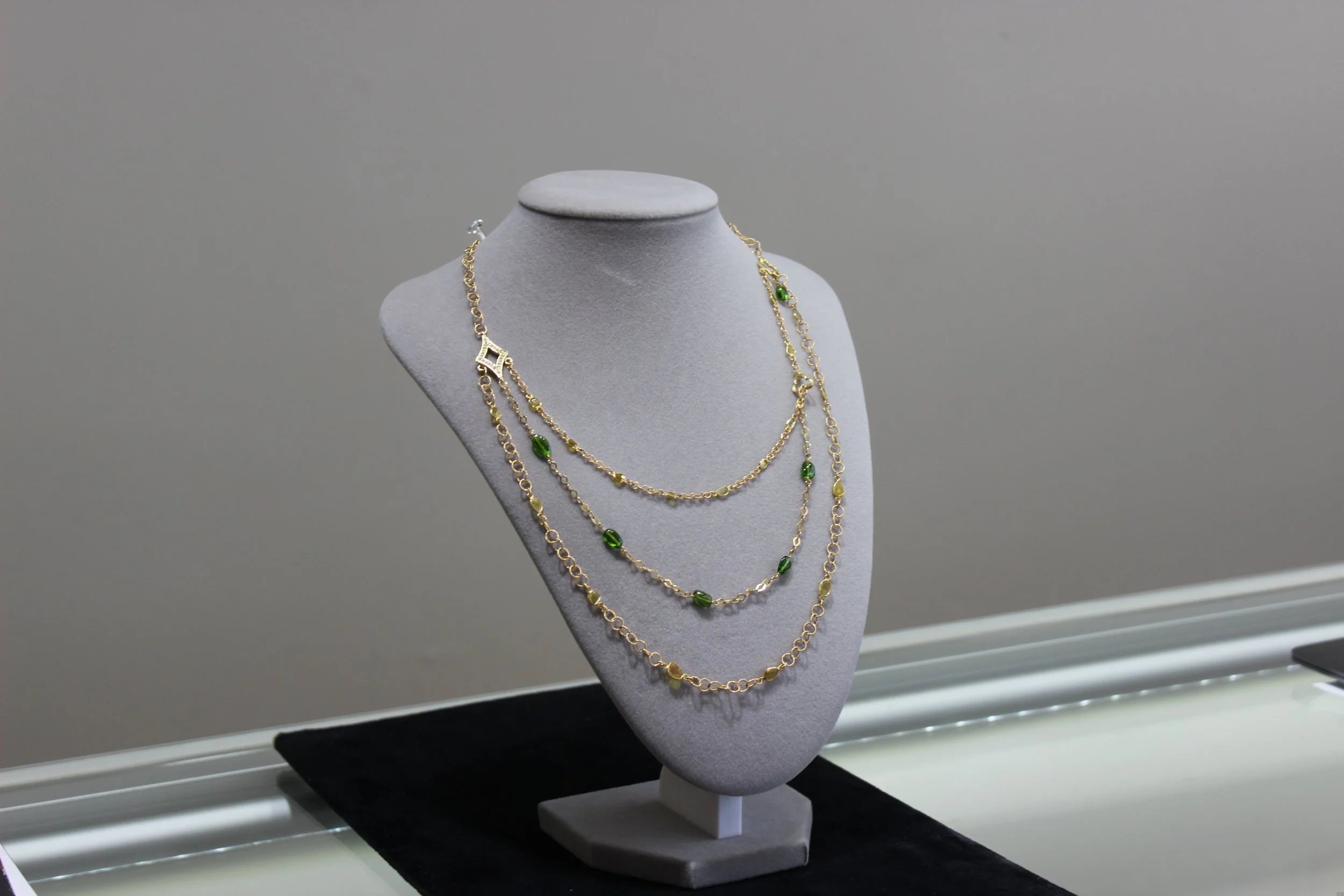 A display of layered gold necklaces with green stones on a grey necklace stand in a jewelry store showcase.