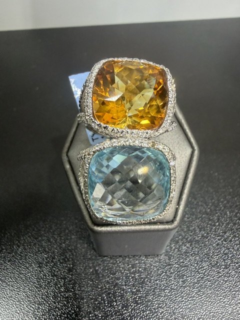 Two large cushion-cut gemstones, an orange-yellow and a pale blue, set in silver rings with diamond accents, displayed on a gray ring holder.