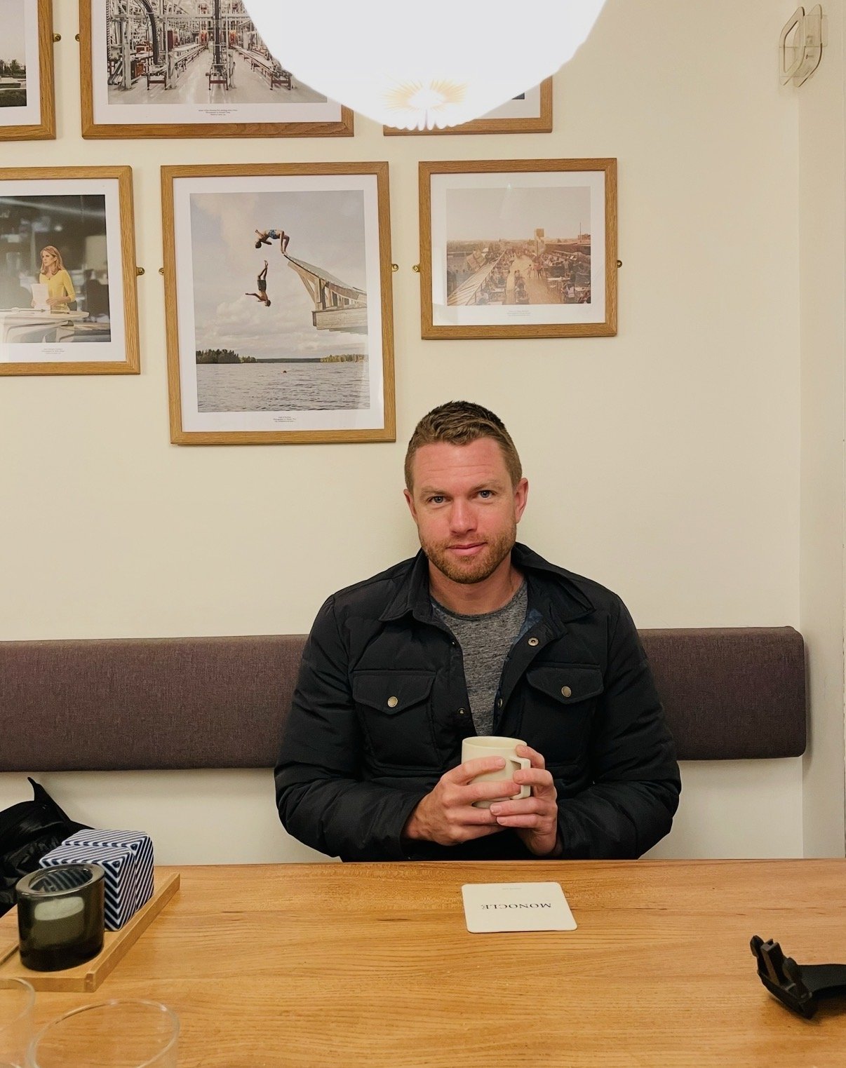 A man with short hair and a beard, wearing a black jacket and gray shirt, sitting at a wooden table in a cafe, holding a white mug. The cafe has framed photographs of lakes, cityscapes, and people on the wall behind him. There are candles and a striped box on the table.