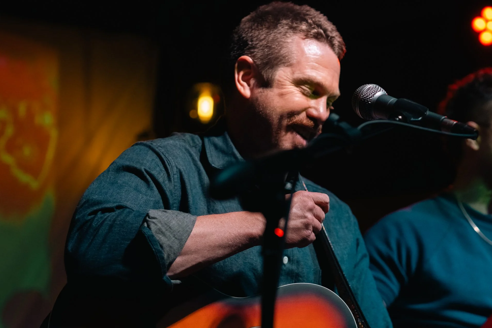 A man with curly hair and a beard smiling while playing an acoustic guitar and singing into a microphone on stage, with warm music venue lighting.