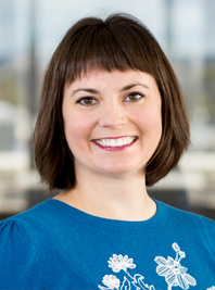 A young woman with short brown hair, smiling, wearing a blue shirt with white floral designs, standing in an office environment with large windows in the background.