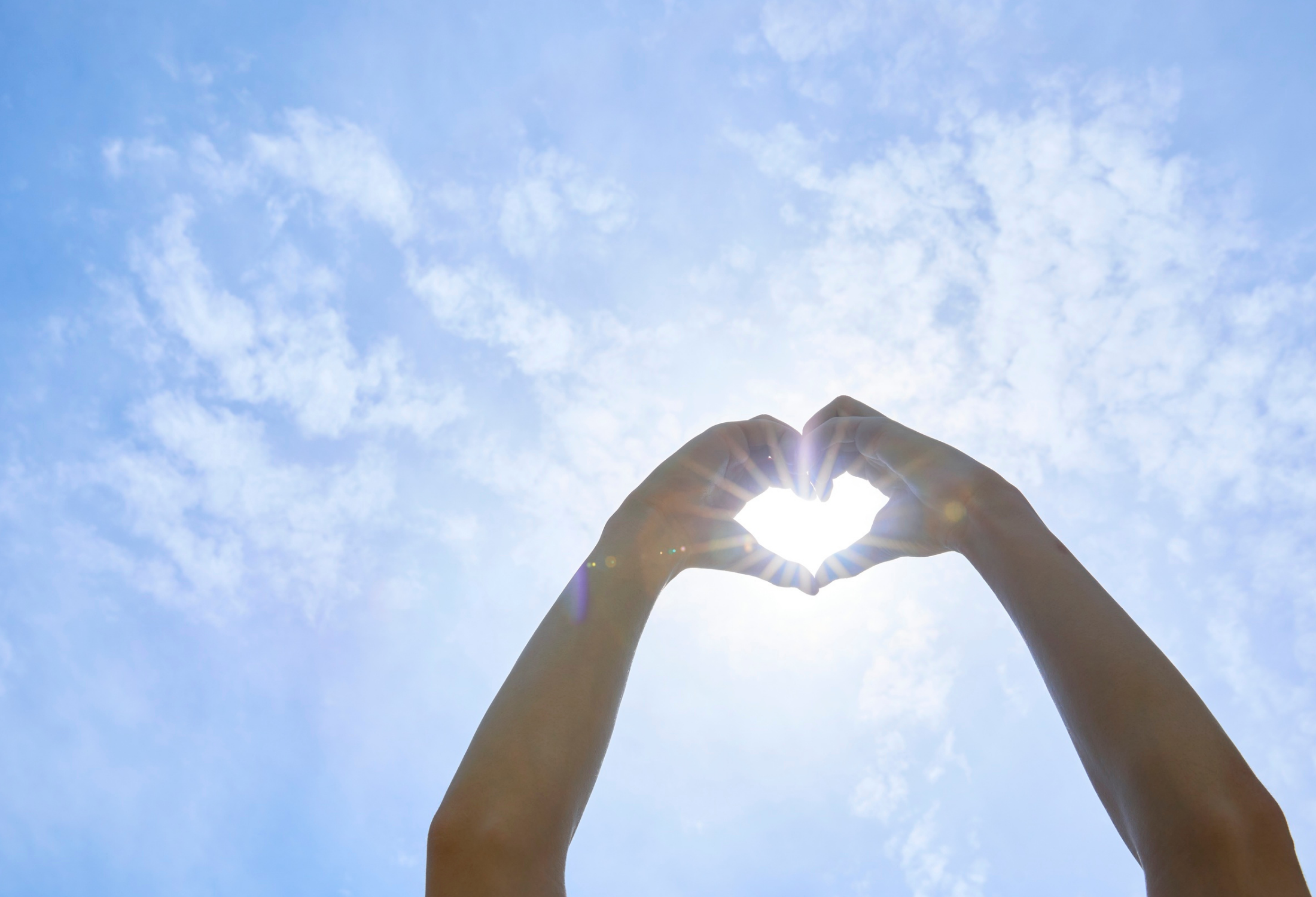 Hands forming a heart shape against a bright blue sky with scattered clouds and a sun glare.