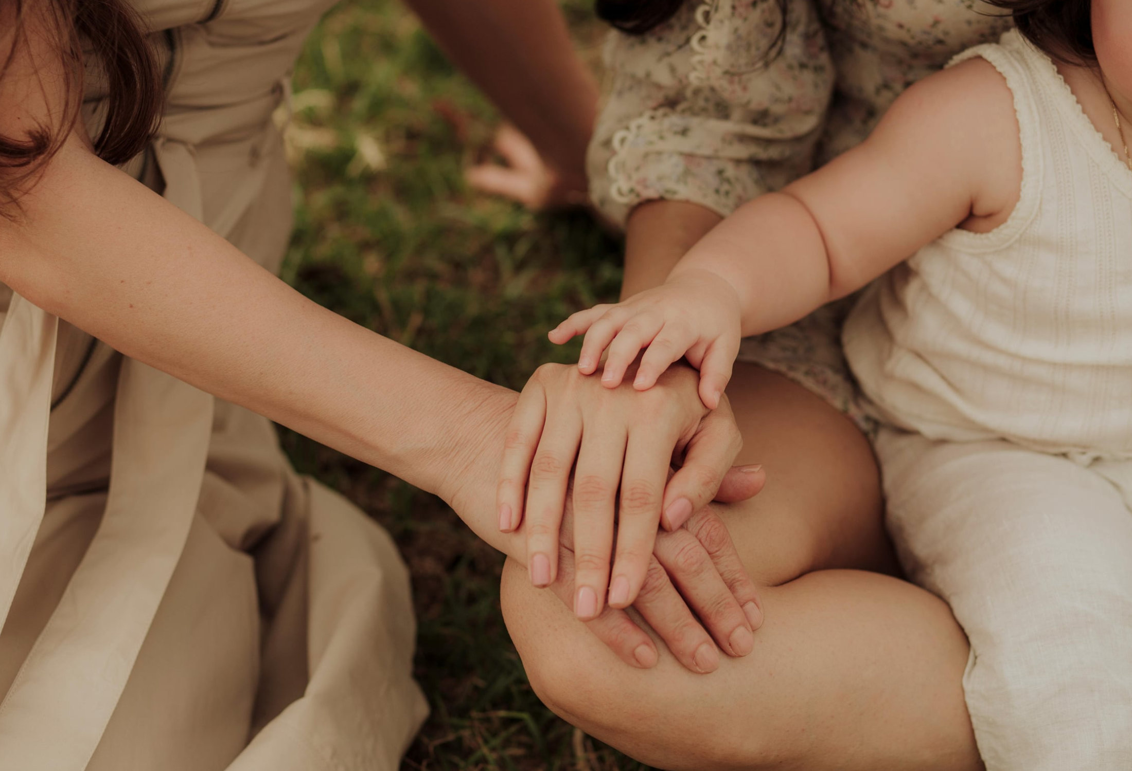 Close-up of children holding hands with an adult, lying outdoors on grass, wearing light-colored clothing.