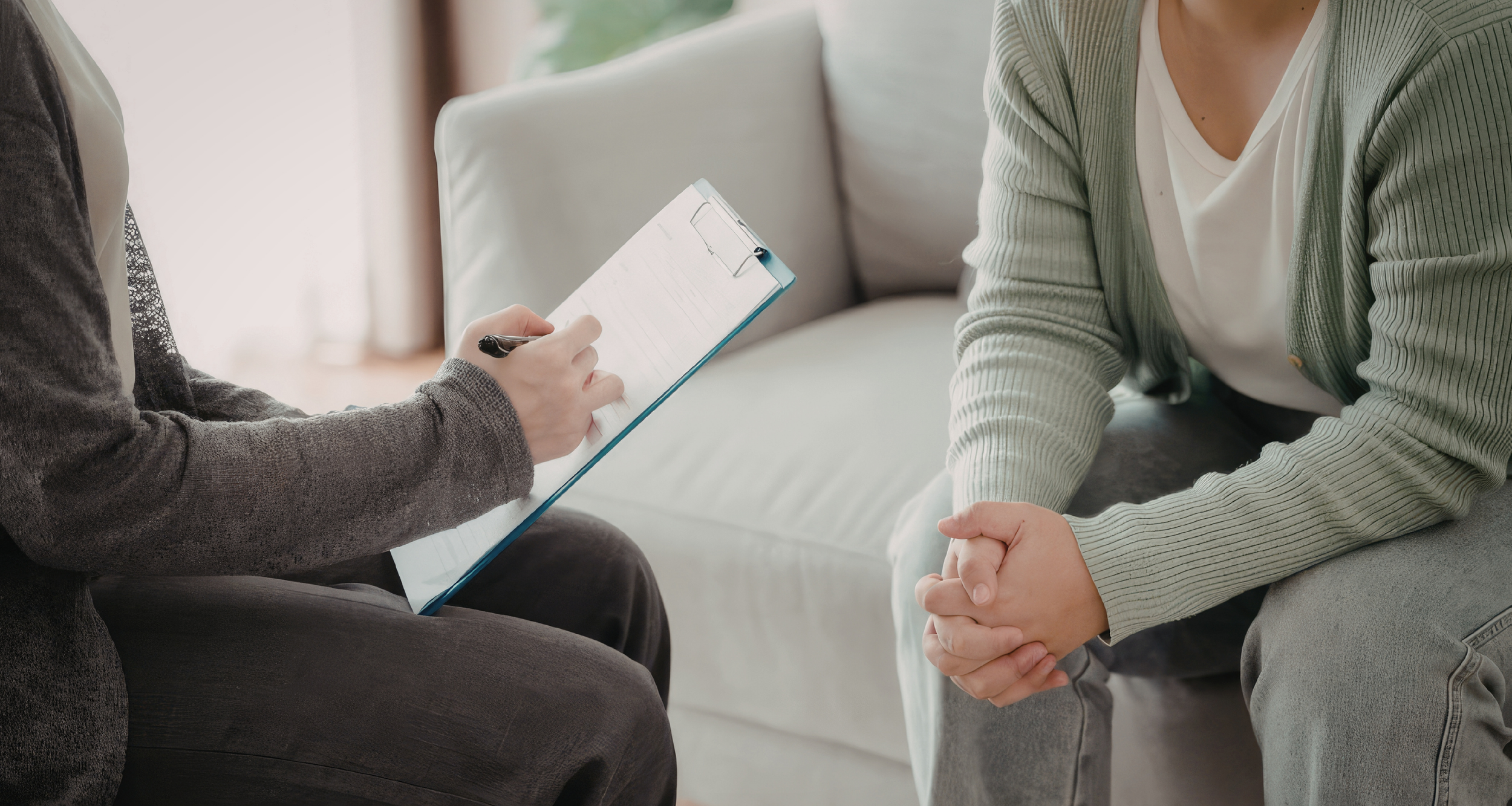A person sitting on a couch holding a clipboard and pen, interviewing or talking to another person who is sitting on the floor with hands clasped together.