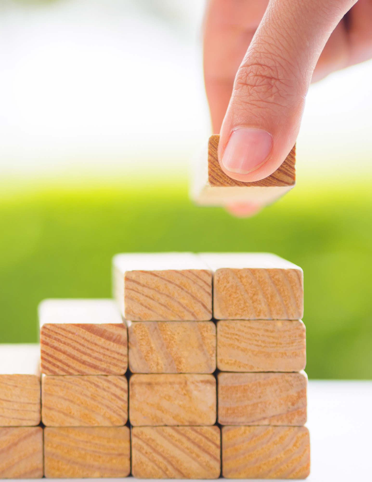 Close-up of a person stacking small wooden blocks with natural wood grain, on a white surface with a blurred green background.