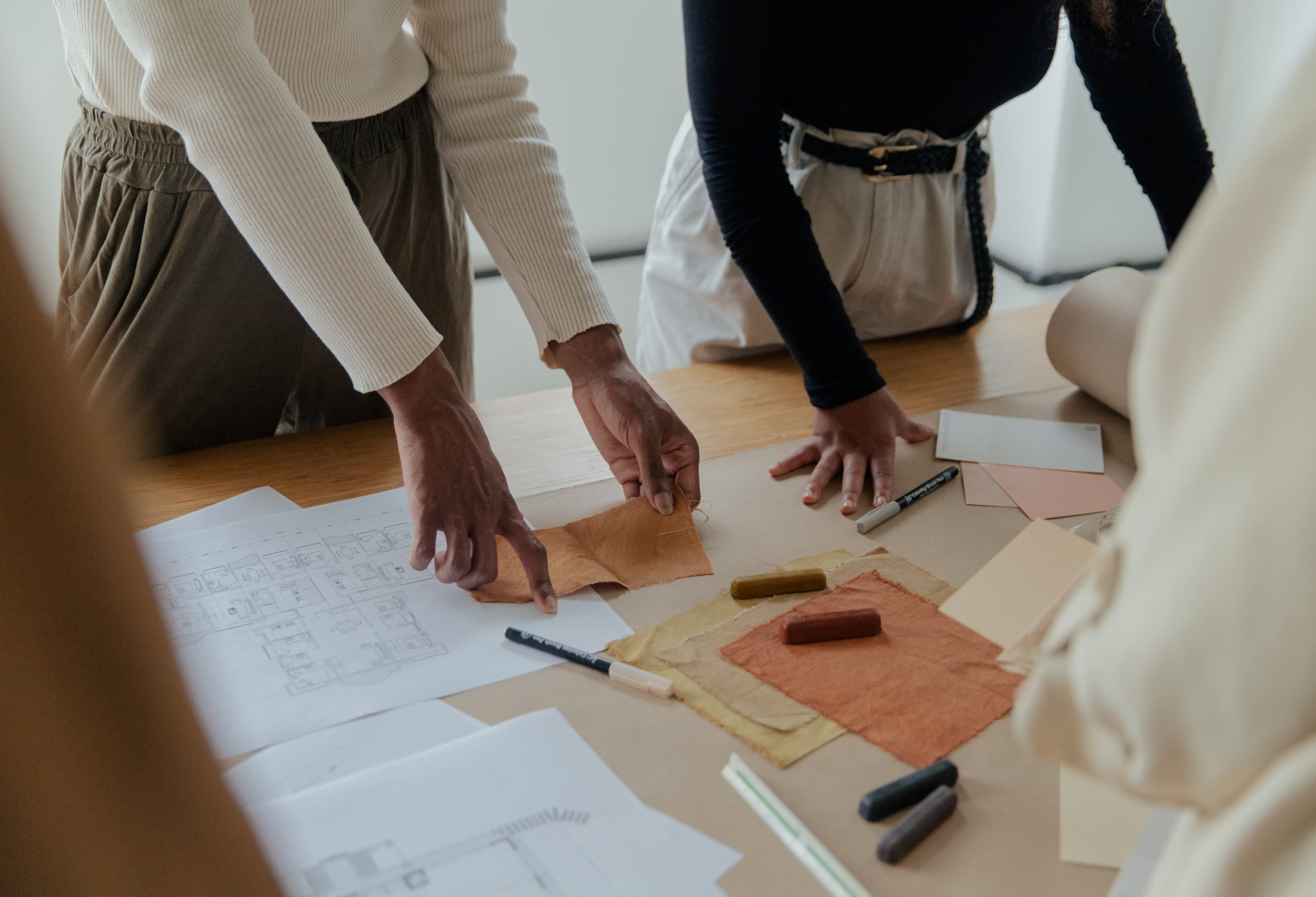 Two people are working on an interior design project, looking at fabric samples, color swatches, and architectural plans laid out on a table.