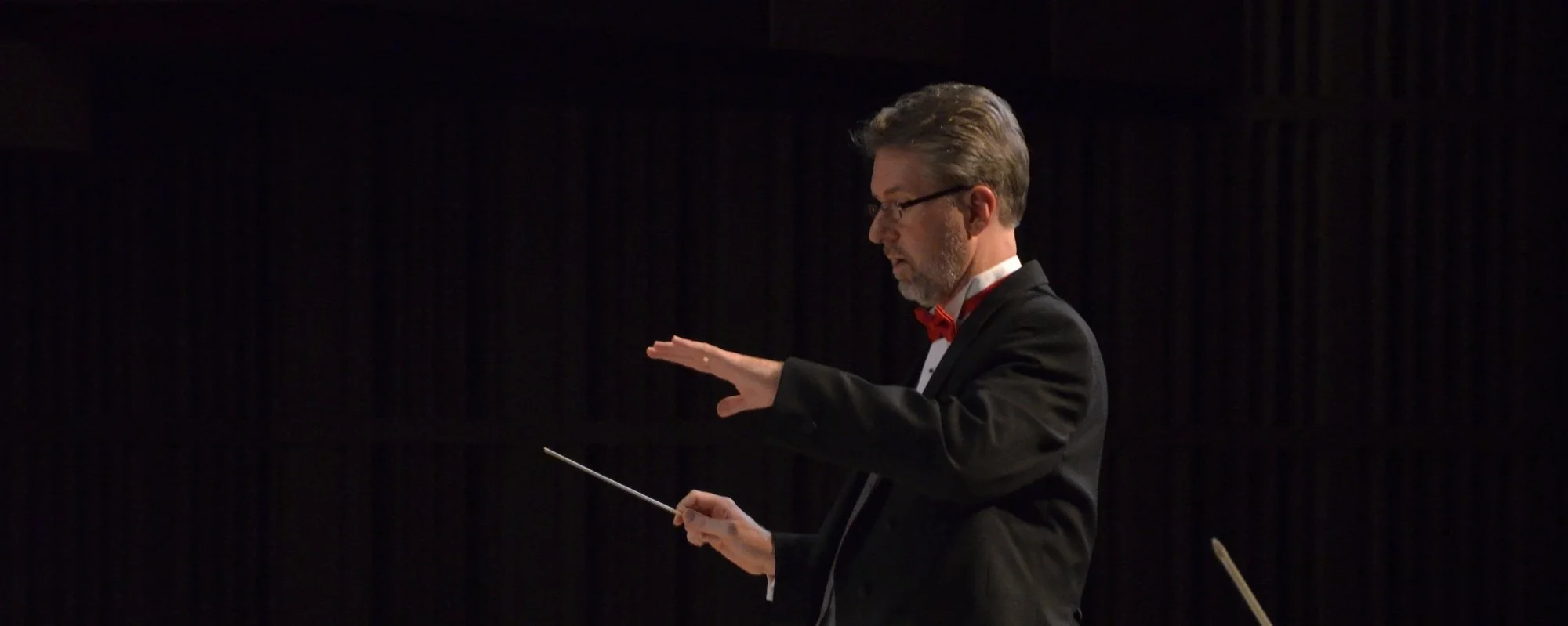 A middle-aged man with gray hair and glasses, dressed in a black tuxedo with a red bow tie, conducting an orchestra in a dark concert hall.