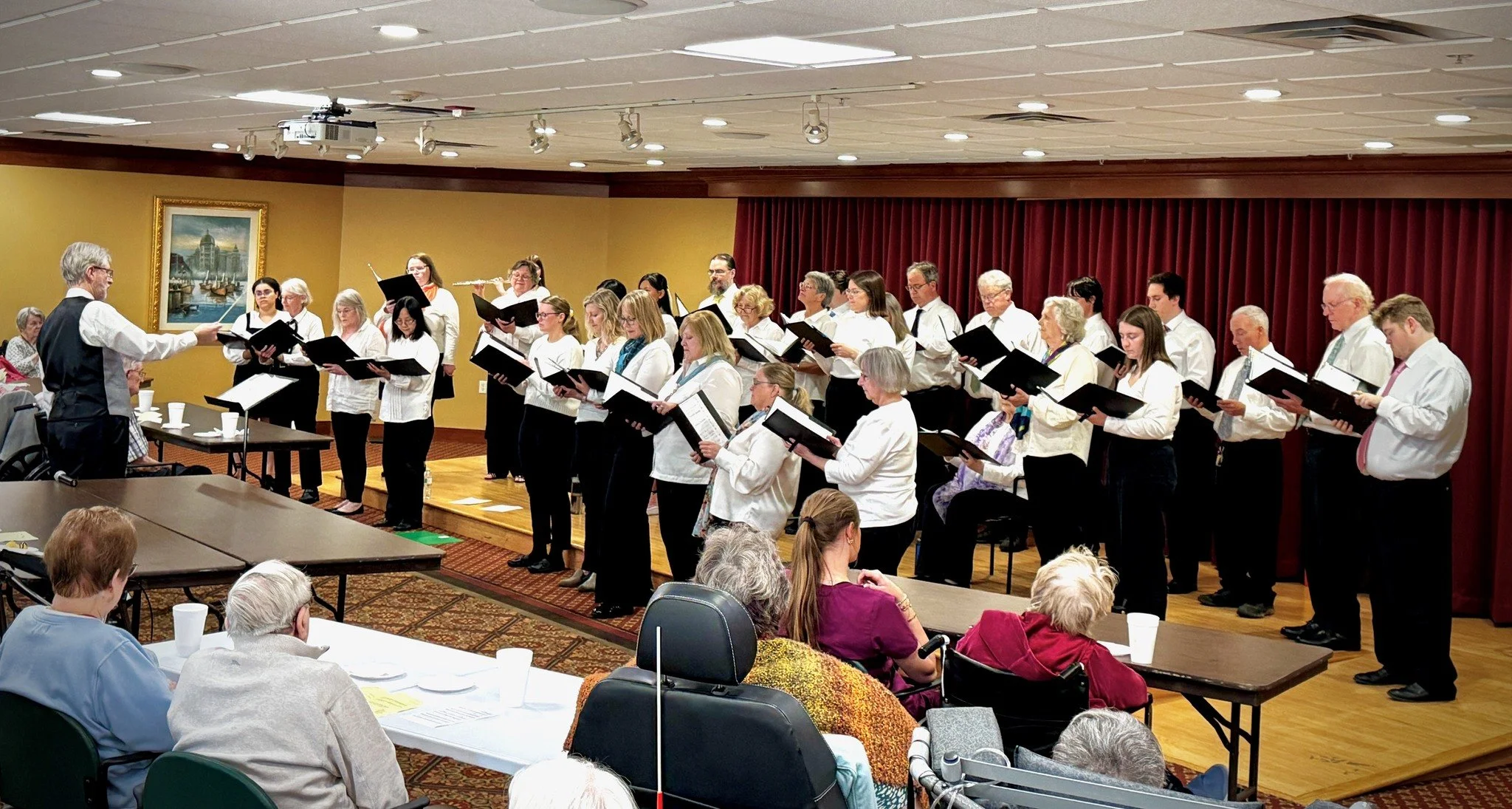 A choir group standing on a stage, led by a conductor, performing in front of an audience in a room with yellow walls and red curtains.