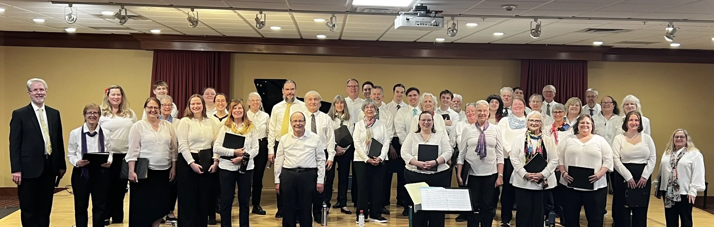 A large group of choir singers standing on stage for a performance, with some holding music folders, under stage lighting in a concert hall.