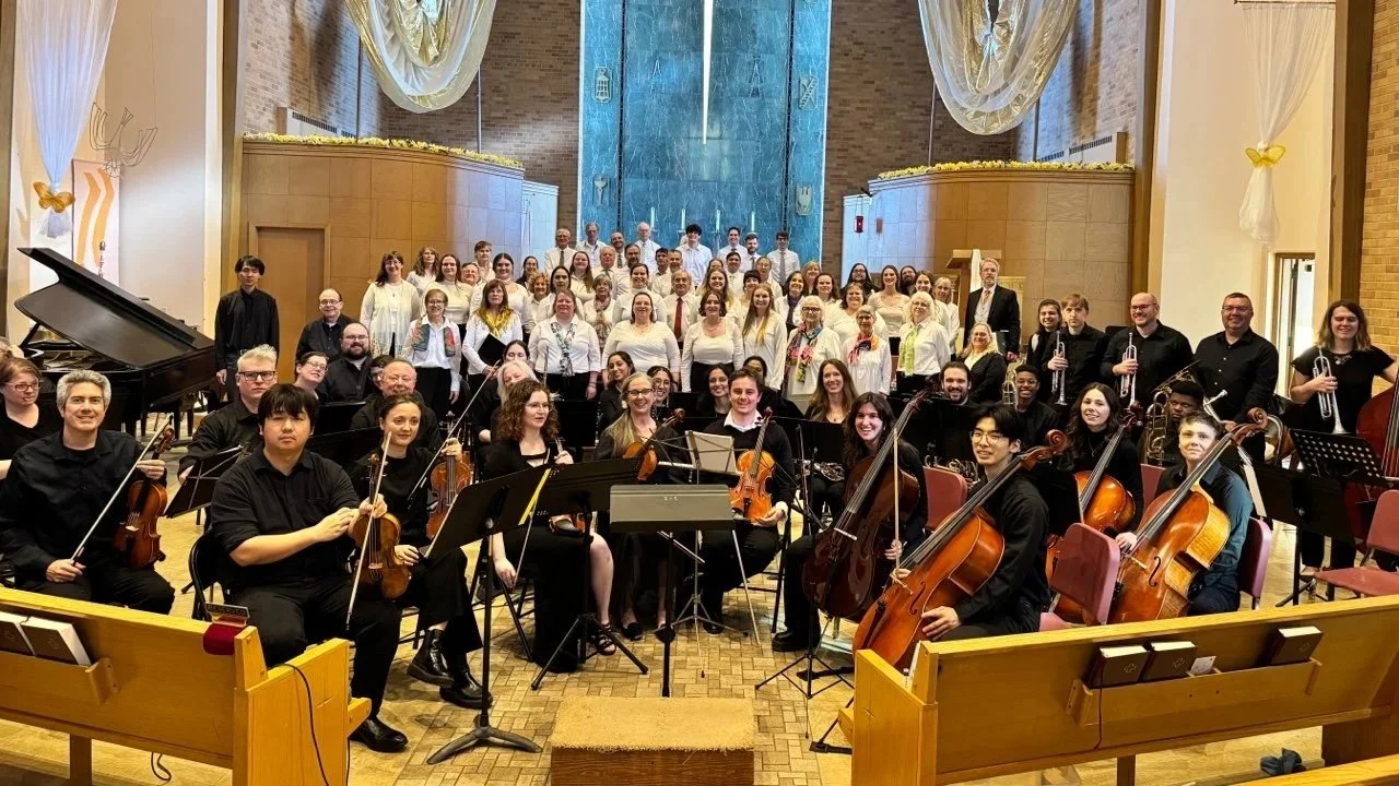 Orchestra and choir group on stage in a concert hall, with string musicians in the front, wind musicians in the middle, and choir singers in the back.