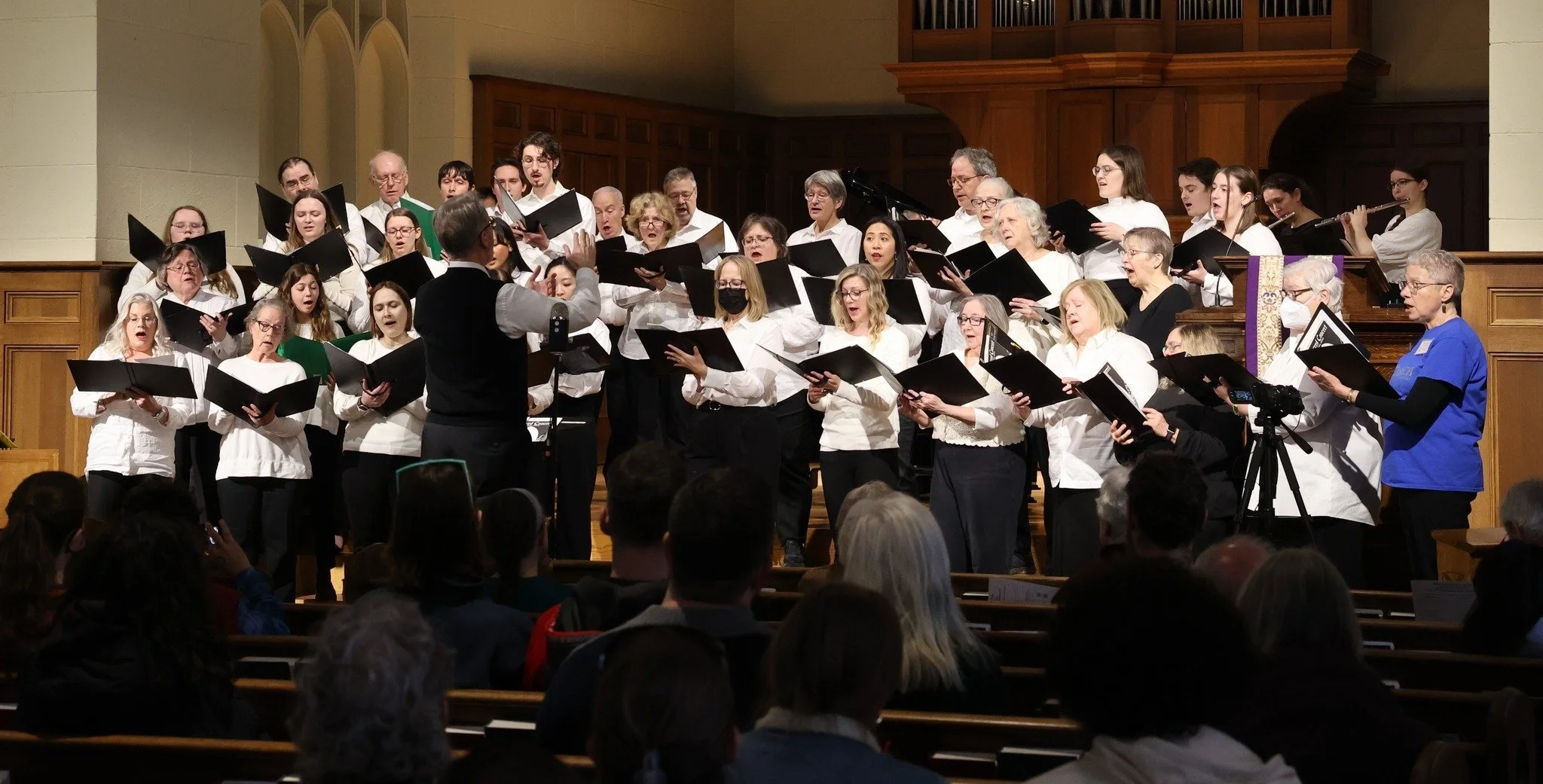 A choir and conductor performing on stage in a church or concert hall, with audience members seated in front.