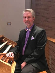 A man with gray hair and a beard, dressed in a black suit, purple shirt, and purple bow tie, sitting at a wooden piano in a room with brick walls.