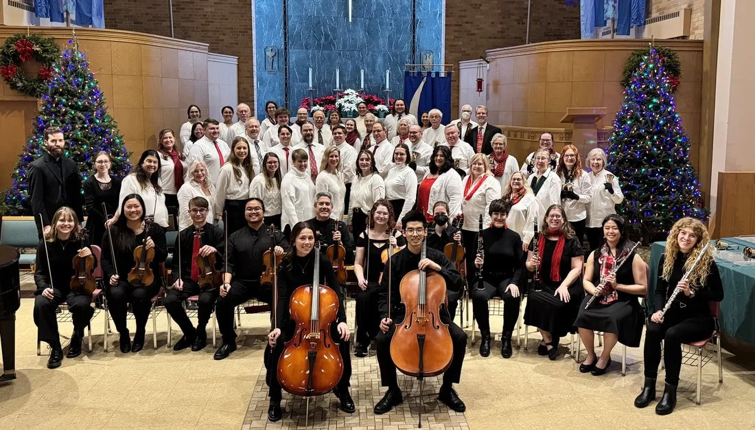 Group photo of a choir and orchestra during a Christmas concert, with decorated Christmas trees on sides and a festive backdrop.