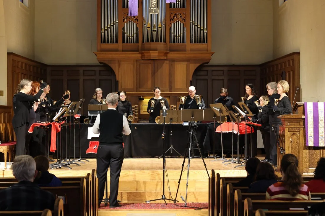 A band performing on stage in a church or concert hall, with a conductor leading, and an audience seated in front.