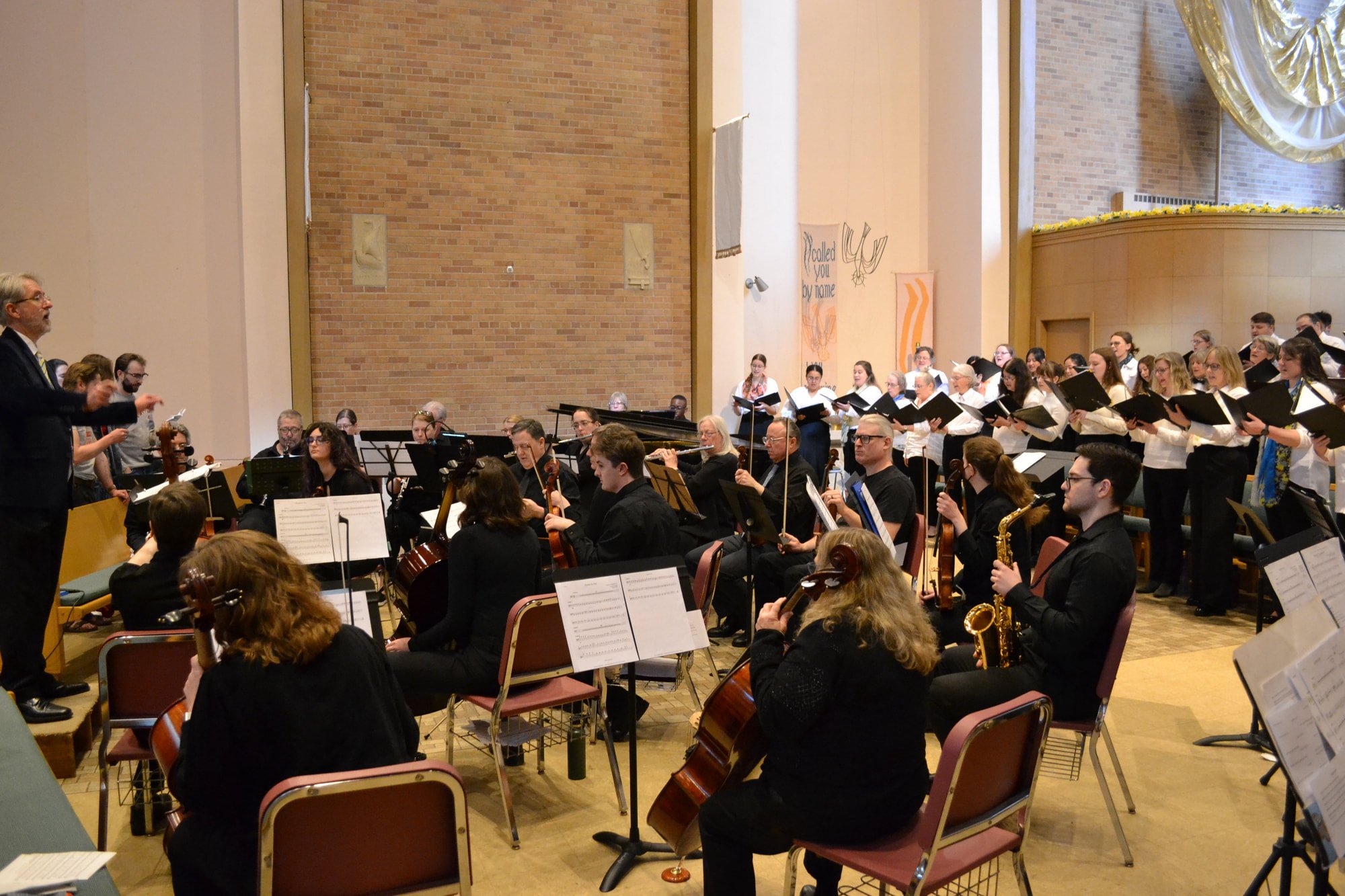 A large choir and orchestra performing in a concert hall with a conductor in front. The background features a brick wall and some banners with religious imagery and words.