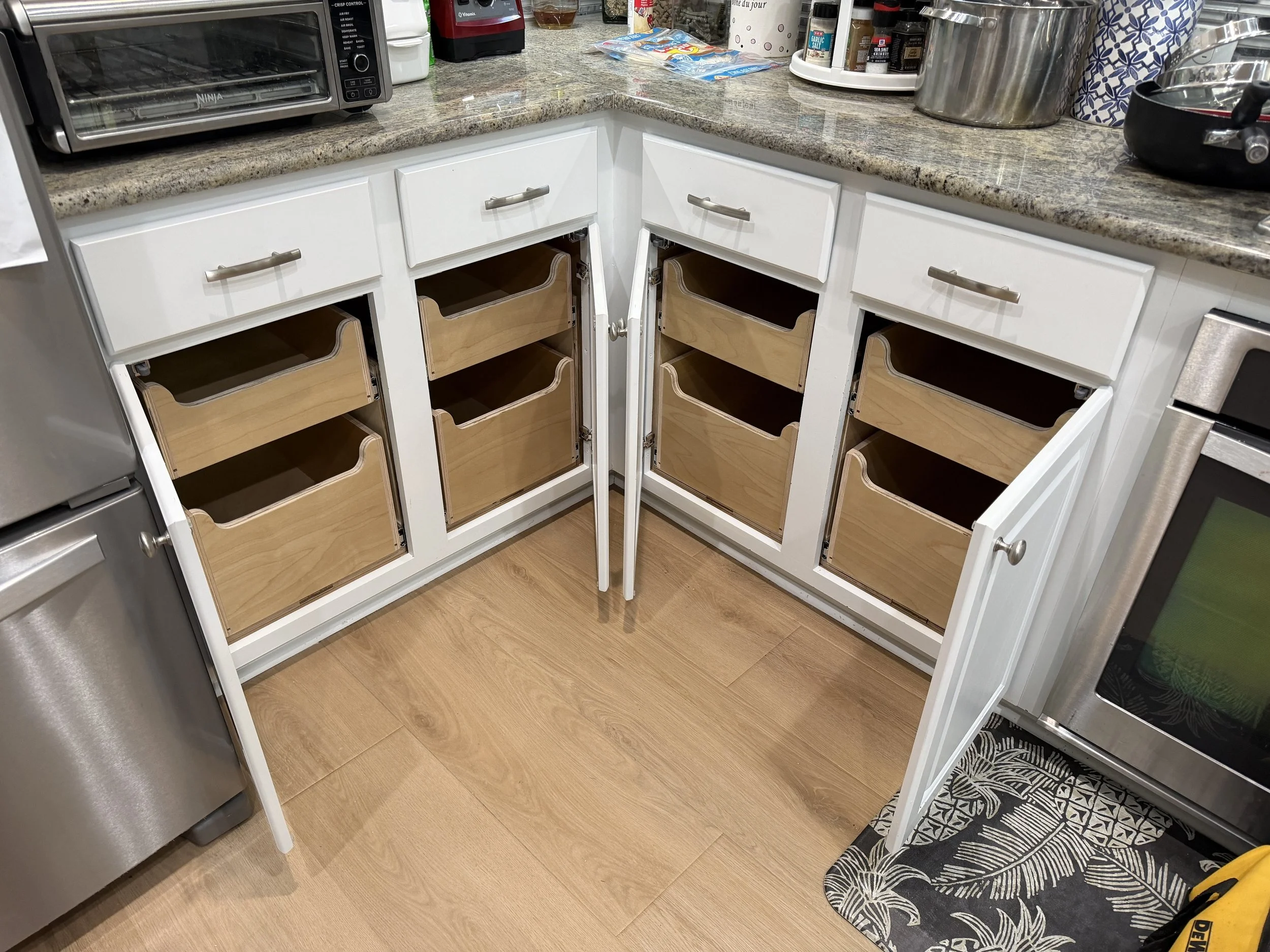 Open kitchen cabinets with drawers and a granite countertop, located next to a stainless steel refrigerator and a stove. The cabinet drawers are made of light-colored wood with a white frame, and some are open showing interior space. The kitchen floo