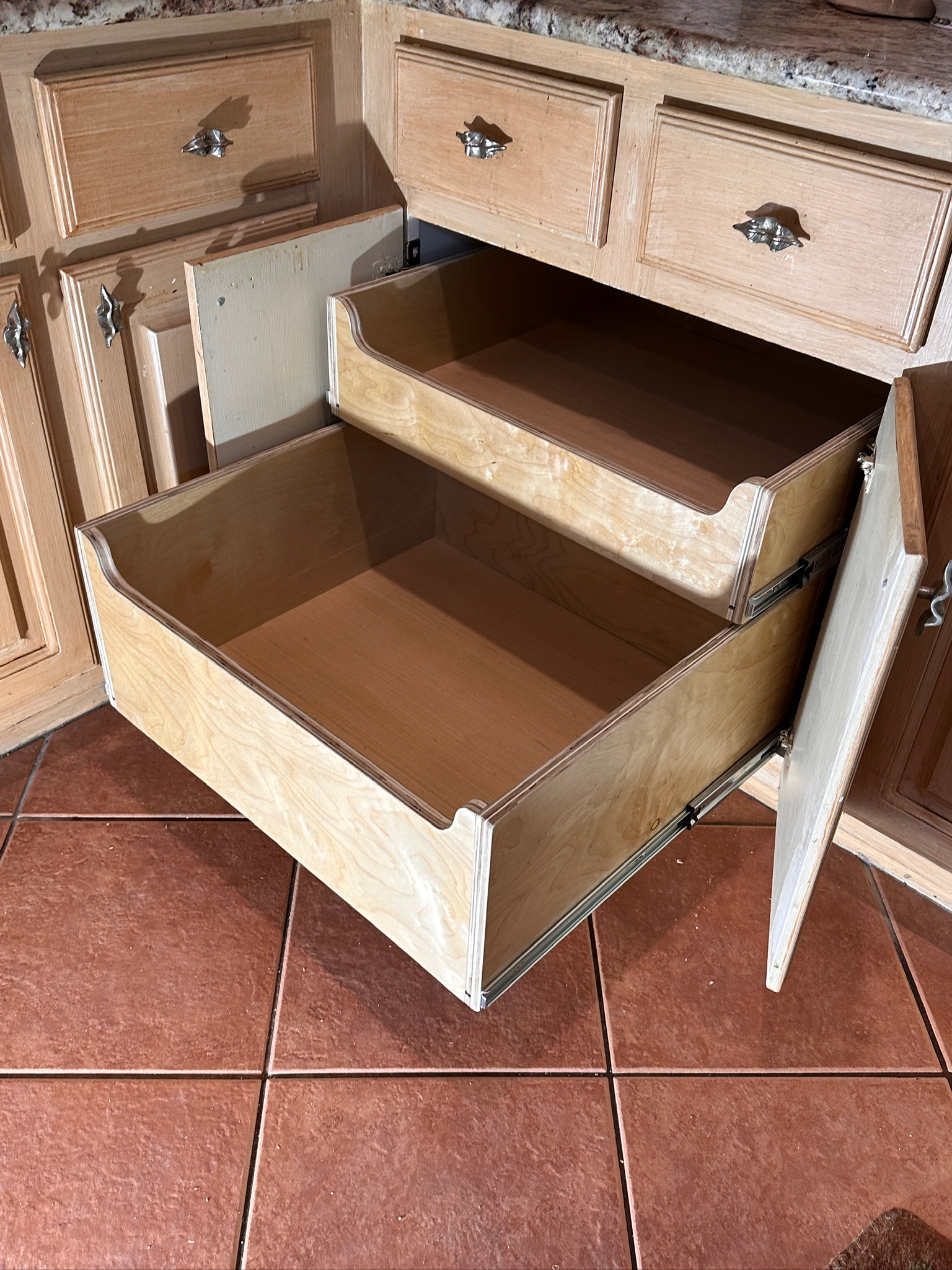 Open kitchen drawer with wooden interior and silver handles, situated beneath a wood cabinet and countertop, placed on reddish-brown tiled floor.
