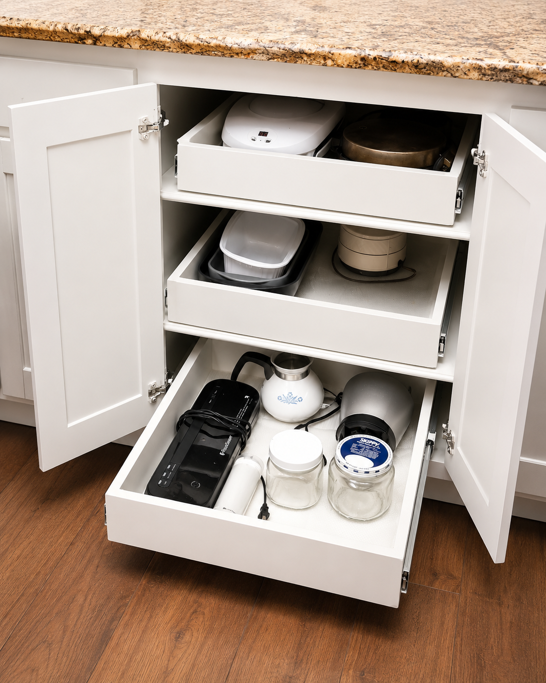 An open white kitchen cabinet with stacked shelves containing various small appliances, kitchenware, and jars, beneath a granite countertop.