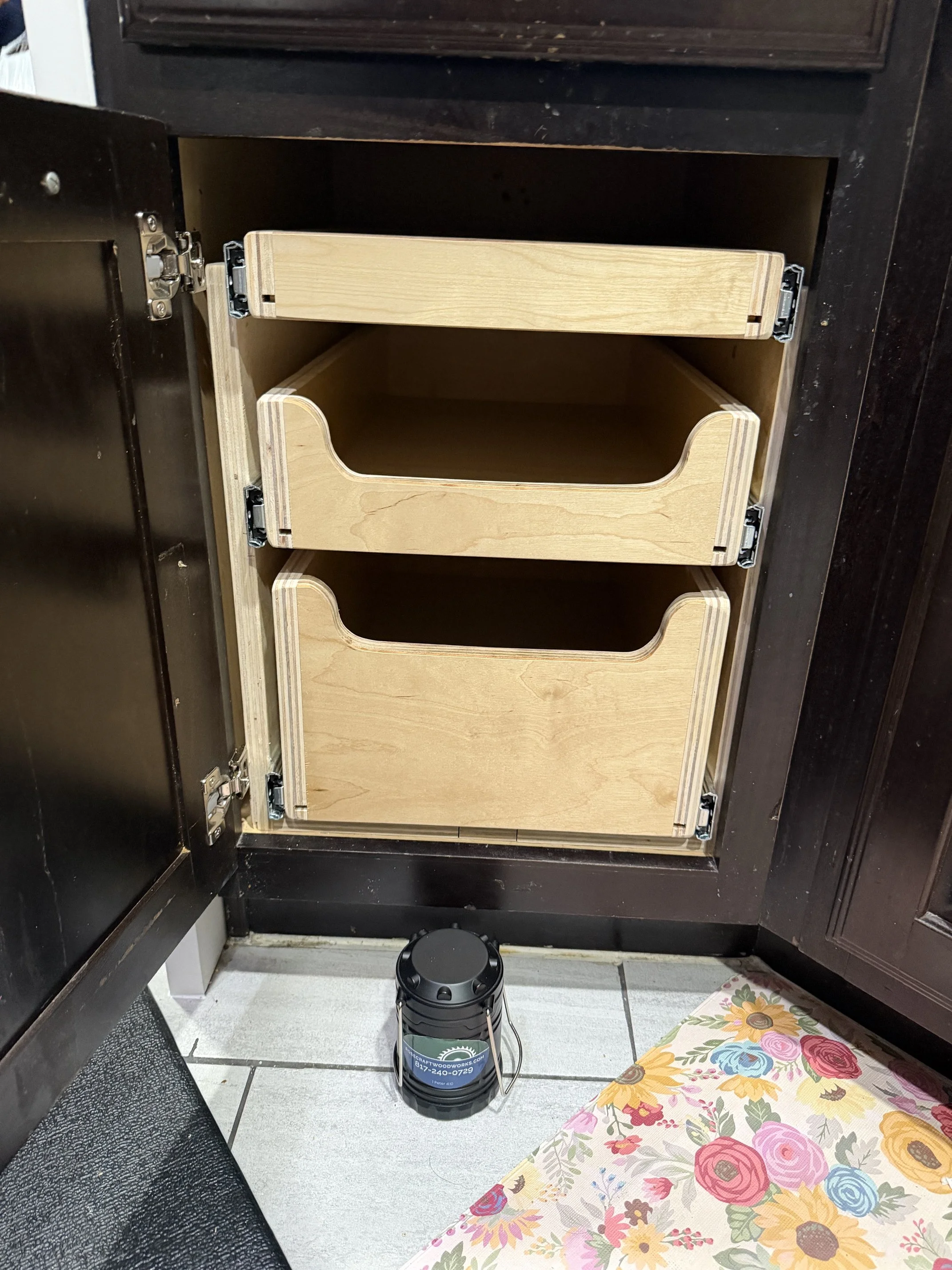 View of an open kitchen cabinet with three wooden pull-out shelves, a lantern with company logo is in front of it.