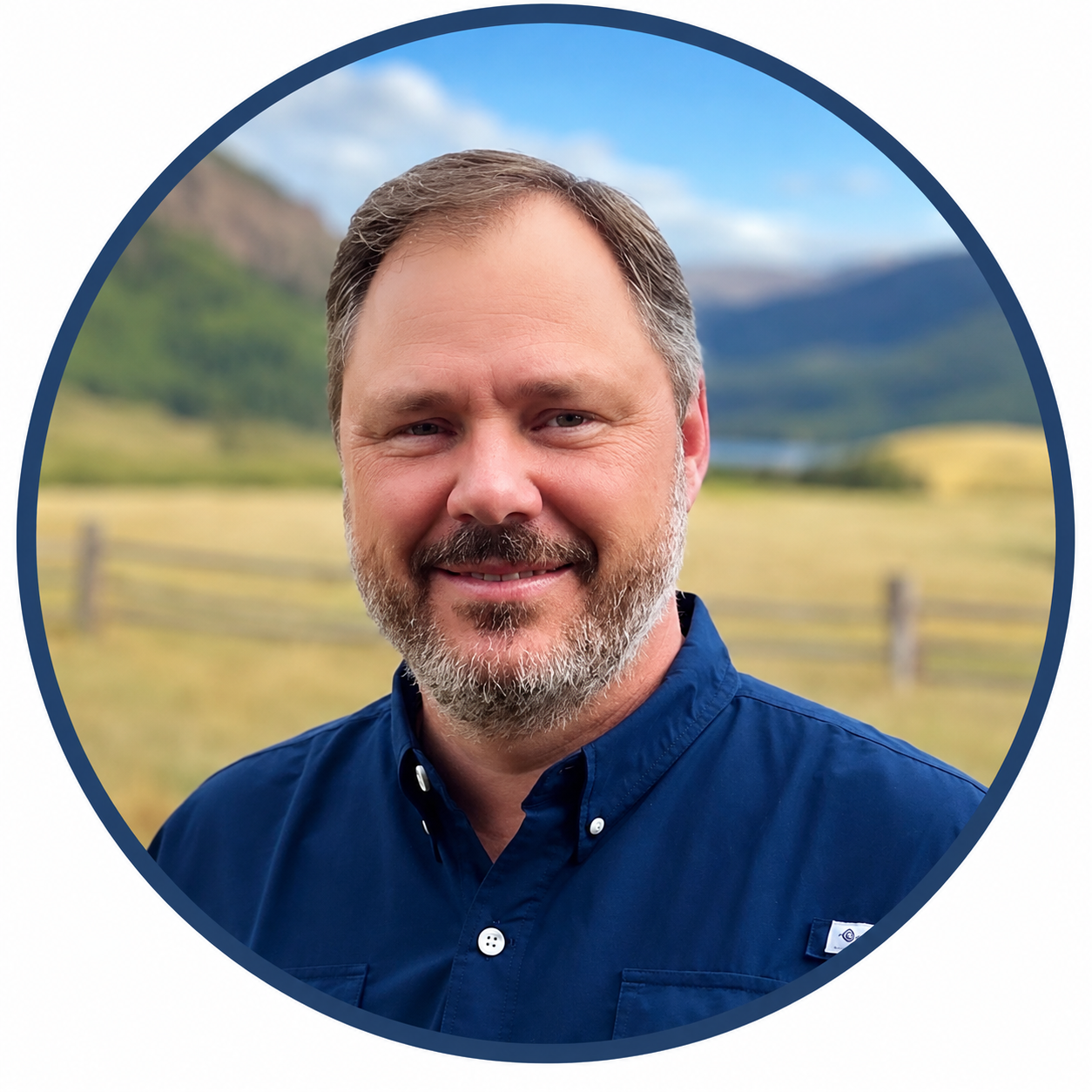 A man with a beard and short hair smiling outdoors with mountains and a field in the background.