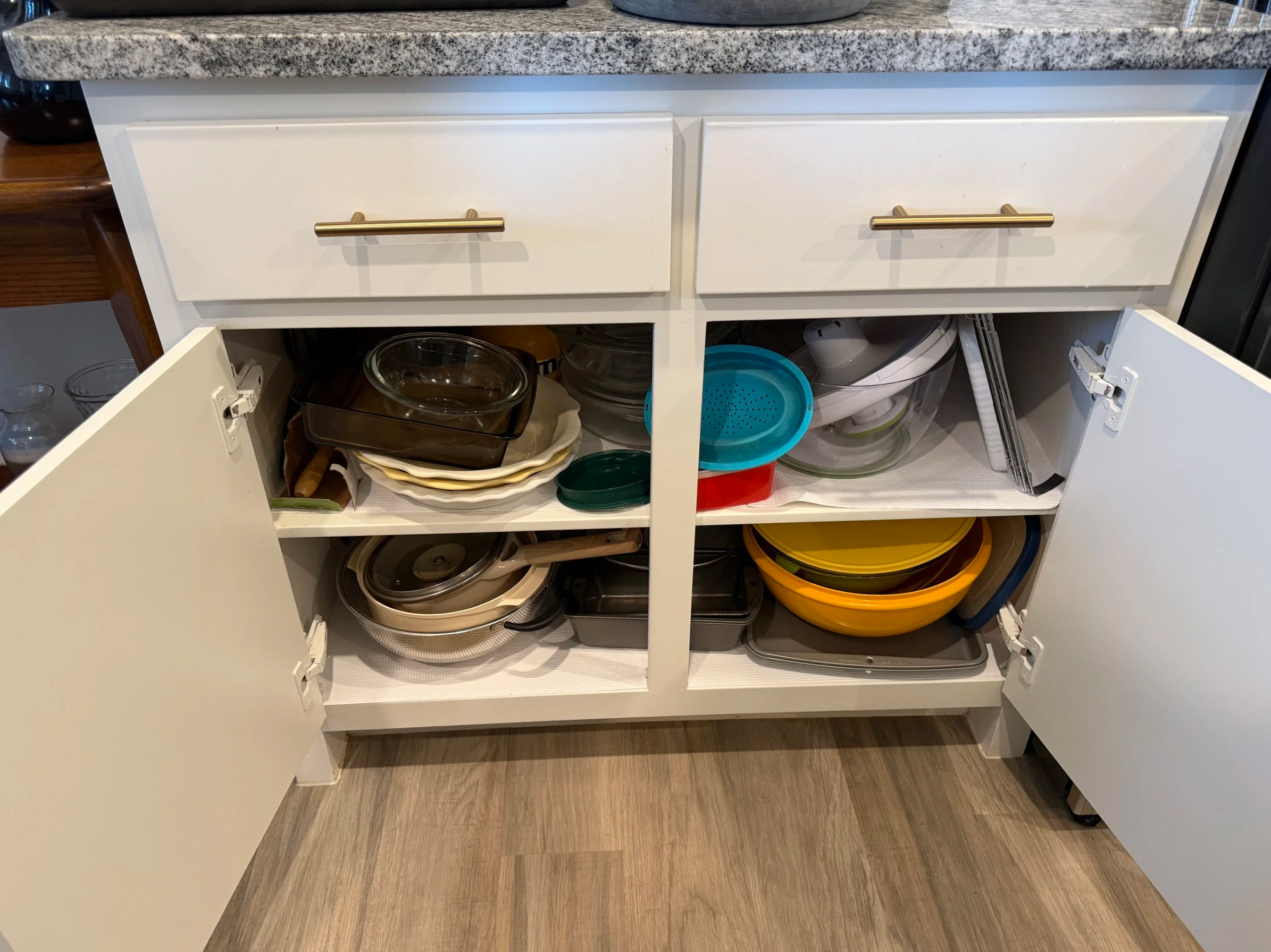 Open kitchen cabinet with various glass and plastic bowls and lids inside, granite countertop above, with hardwood floor beneath.