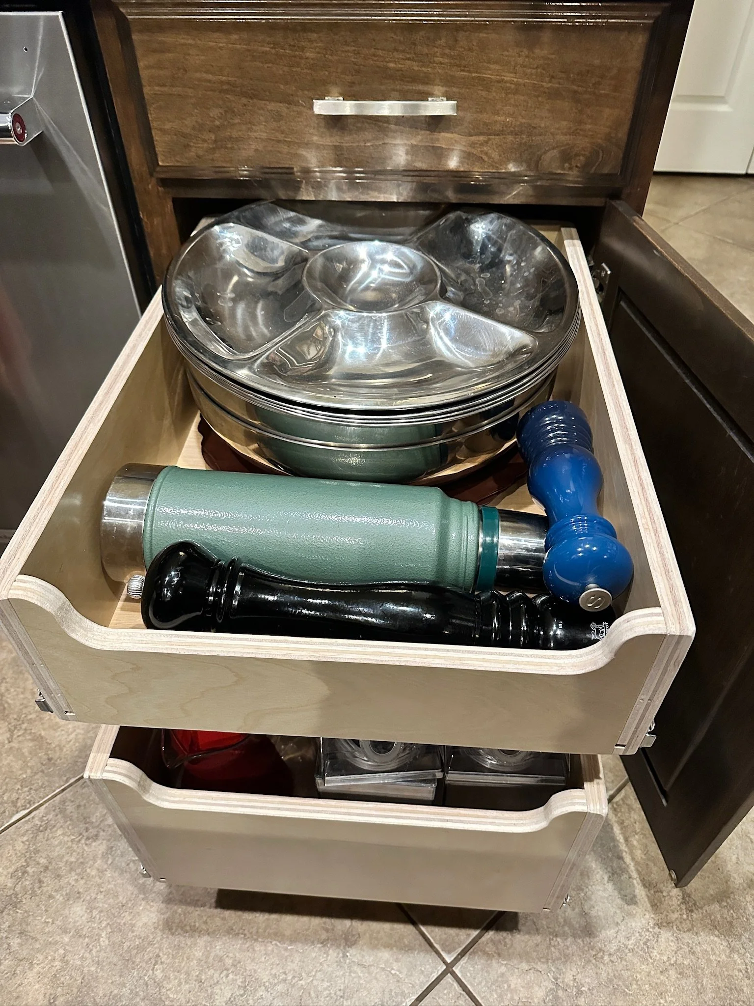 A wooden drawer filled with metal bowls, a green water bottle, black and blue water bottles, all arranged inside a cabinet.
