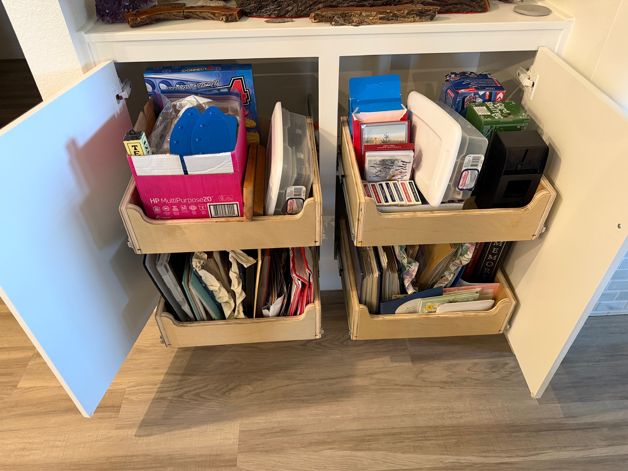 A white open bookshelf with various books, board games, and folders organized inside. The bookshelf is placed against a wall with a wooden floor in front.