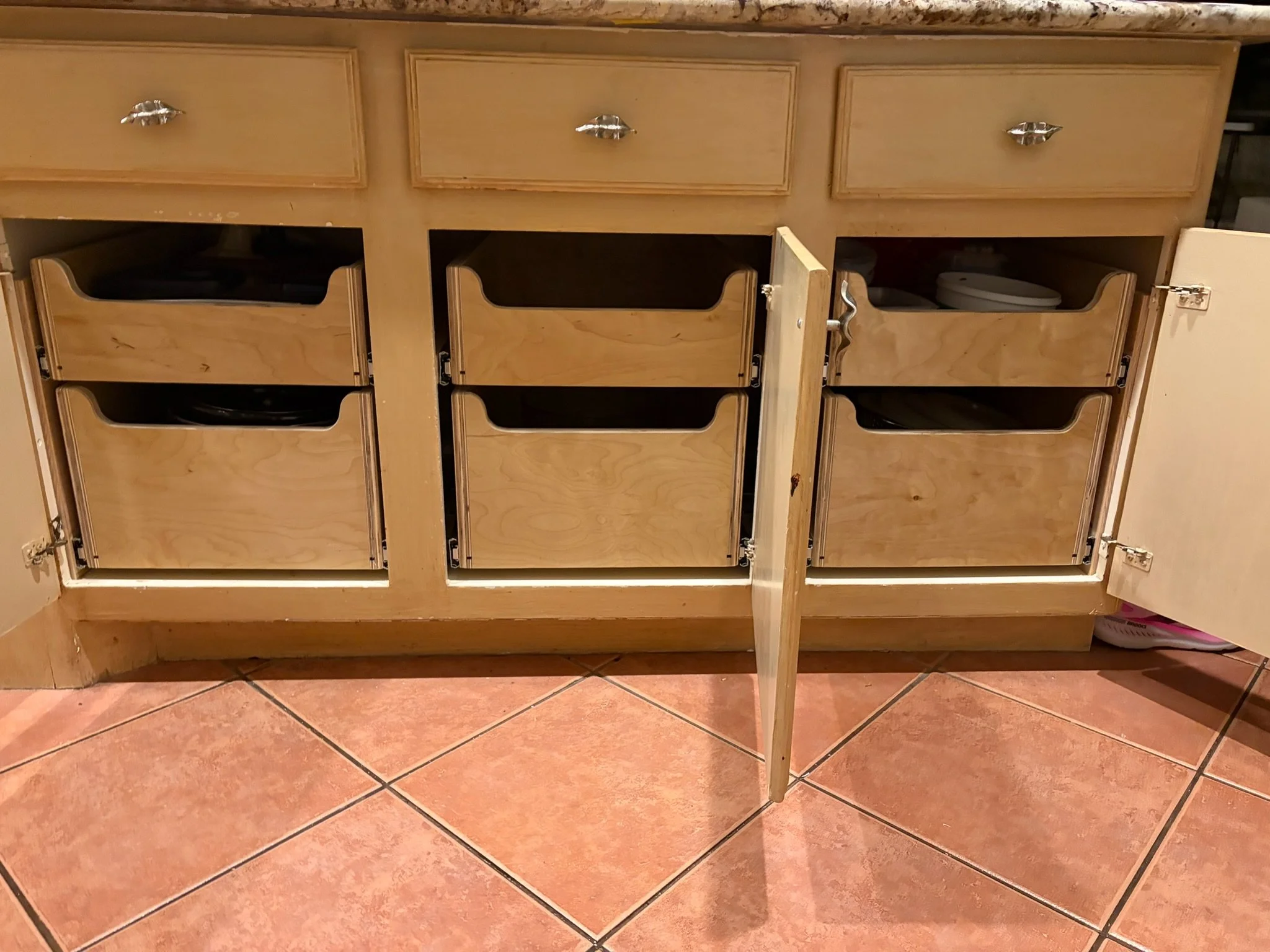 Open kitchen cabinet with three drawers and three doors, with some items visible inside, on a tiled kitchen floor.