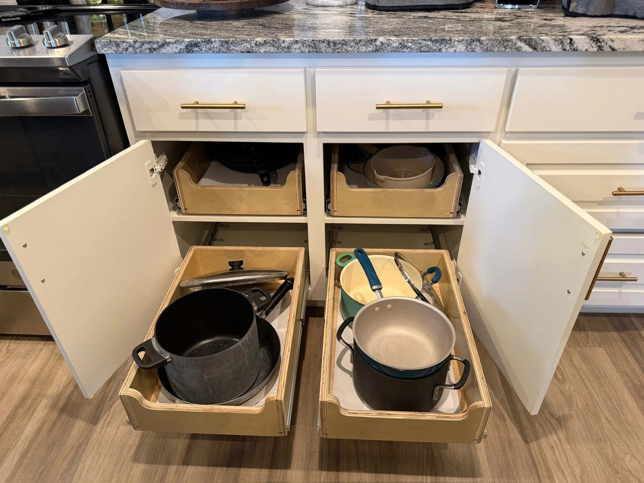 Open kitchen cabinets with cookware and kitchen utensils inside, located under a granite countertop.