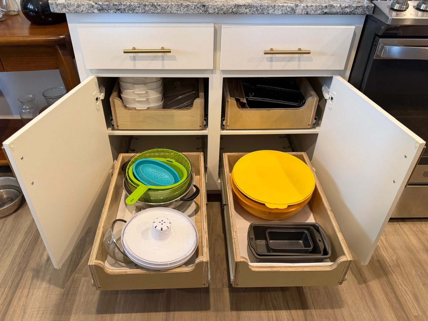 Open kitchen cabinet with dishes, bowls, and containers. Inside the cabinet, there are stacked white dishes, a few baking sheets, mixing bowls, and plastic containers. In front of the cabinets, there are two pull-out drawers containing colorful sieves, bowls, and storage containers.