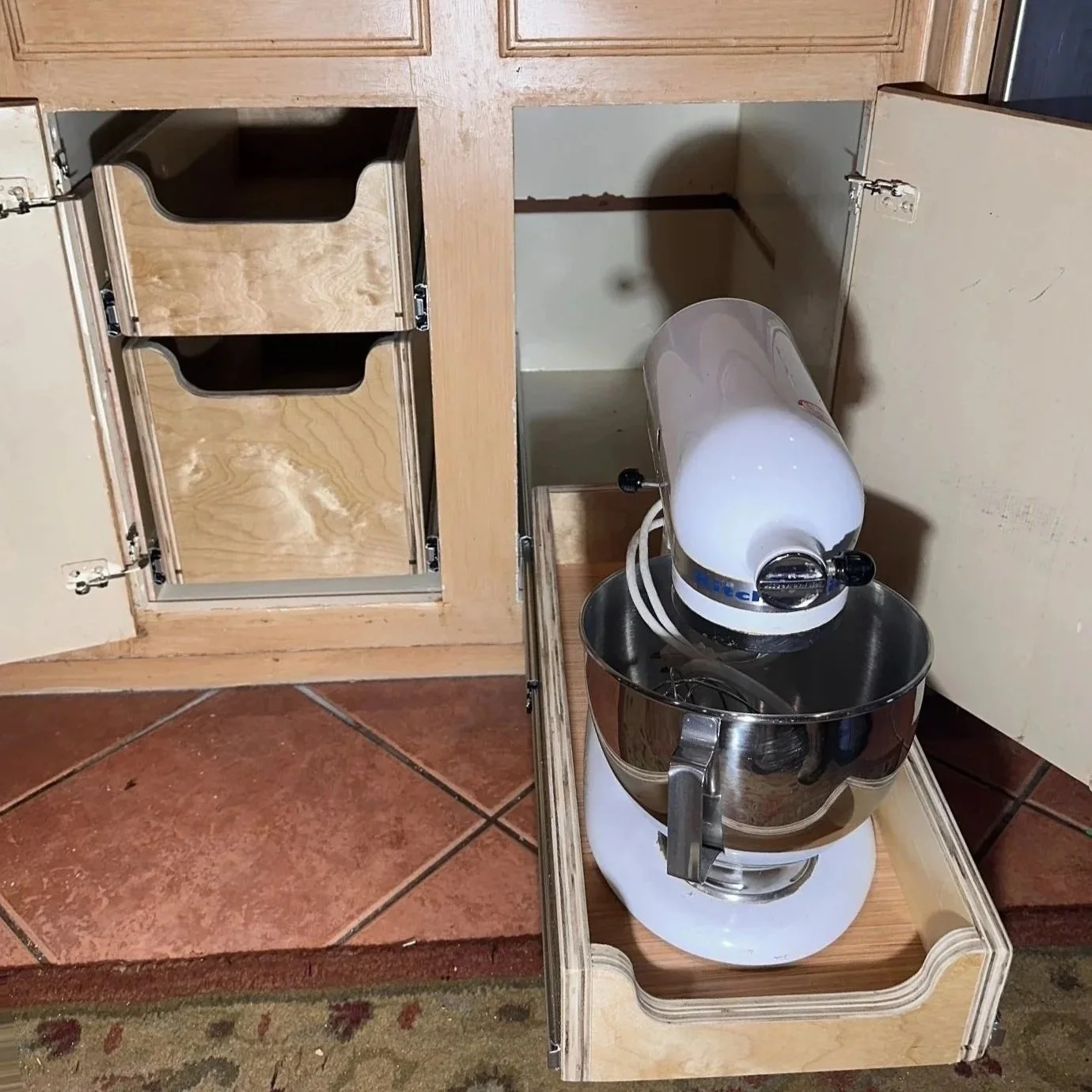Open kitchen cabinet with empty shelves and a stand mixer on a wooden cart in front of the cabinet, on a tiled floor.