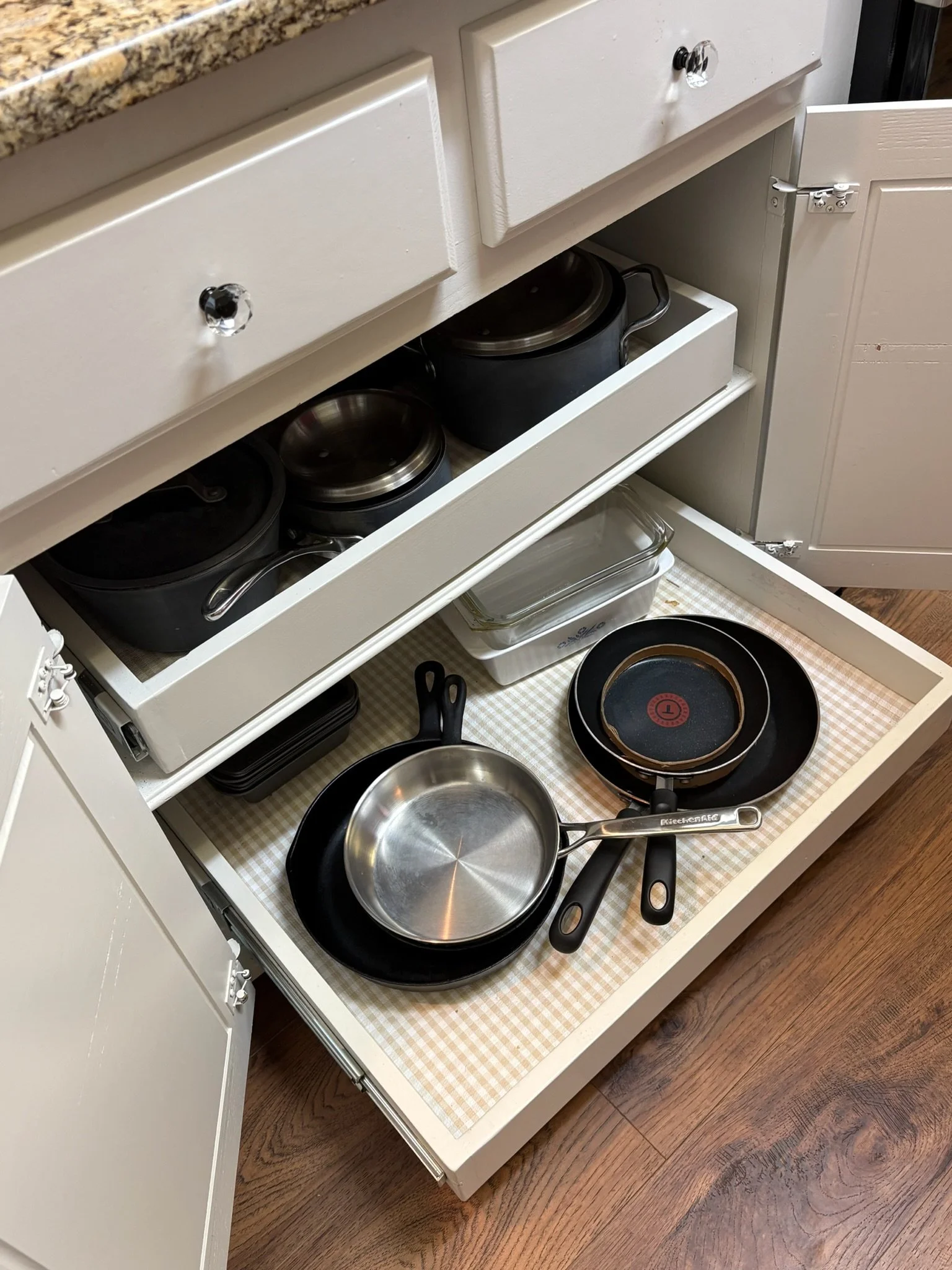 Open kitchen drawer with various pots, pans, and kitchen utensils inside, on a wooden floor.