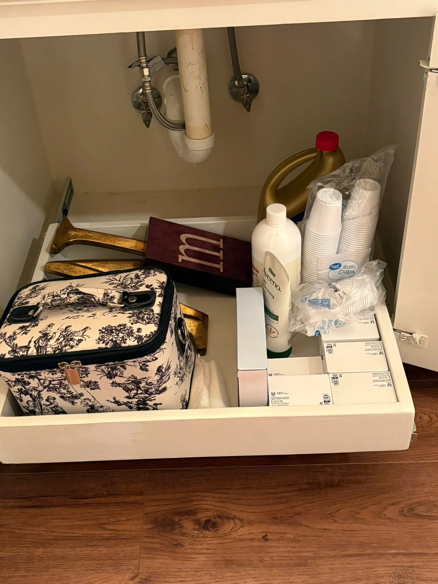 Open kitchen cabinet with plumbing pipes, cleaning supplies, disposable cups, and boxes of medicine or medical supplies, with a patterned bag and a small upholstered stool inside.