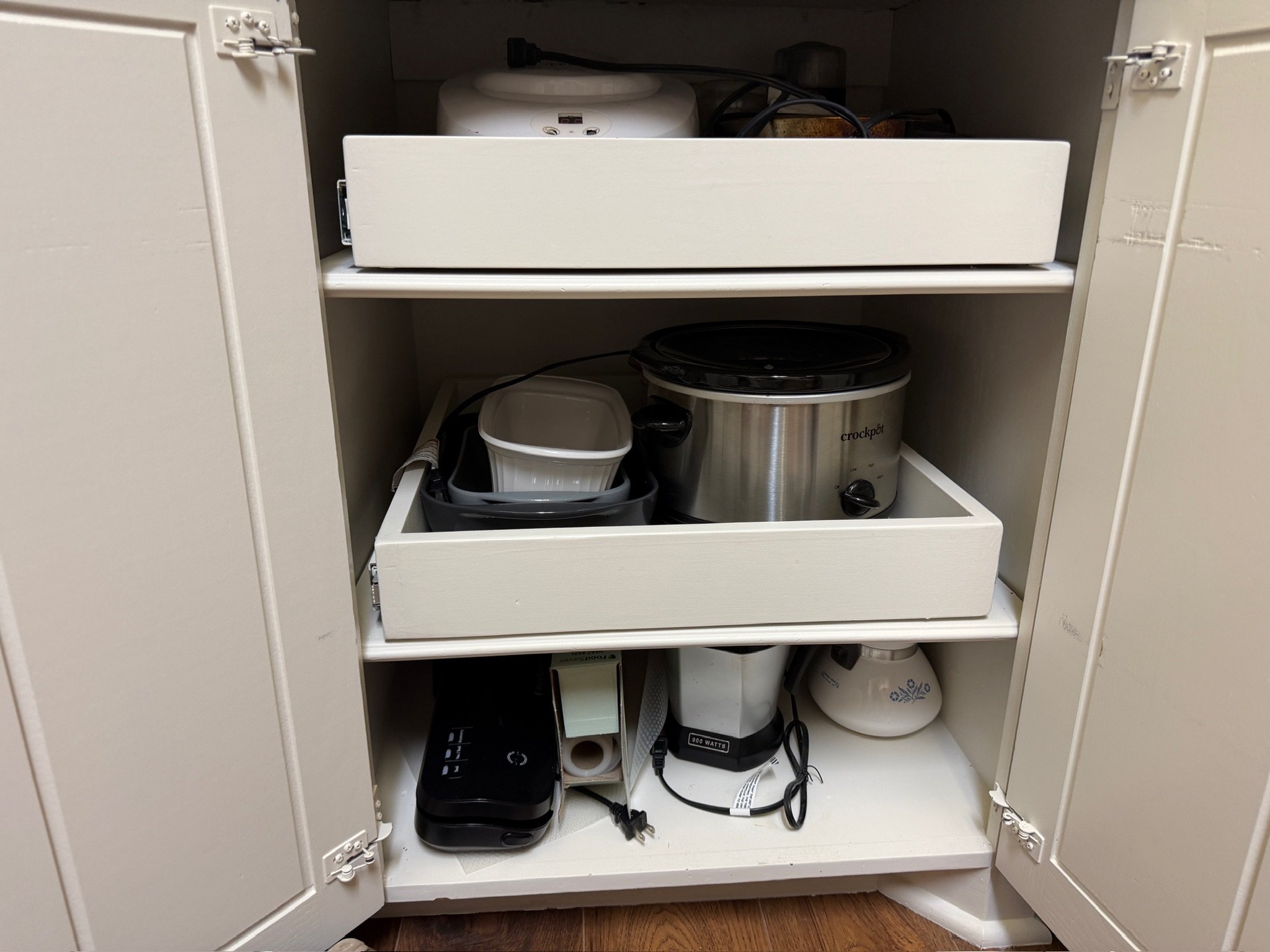 Open kitchen cabinet with appliances including a small rice cooker, a slow cooker, a toaster oven, and various cords and kitchen items stored inside.