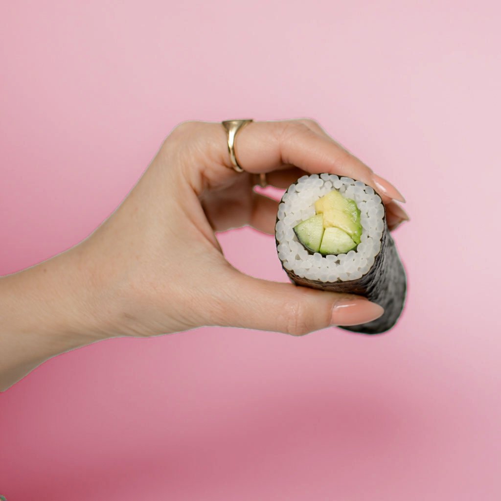 Close-up of a hand holding a piece of sushi with cucumber and yellow filling against a pink background.