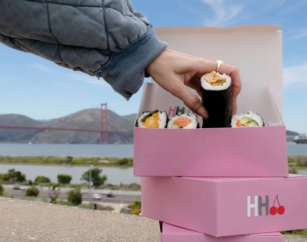 A hand with a gray jacket reaching into a pink box of assorted sushi rolls, with Golden Gate Bridge in the background and water, hills, and a few trees visible.