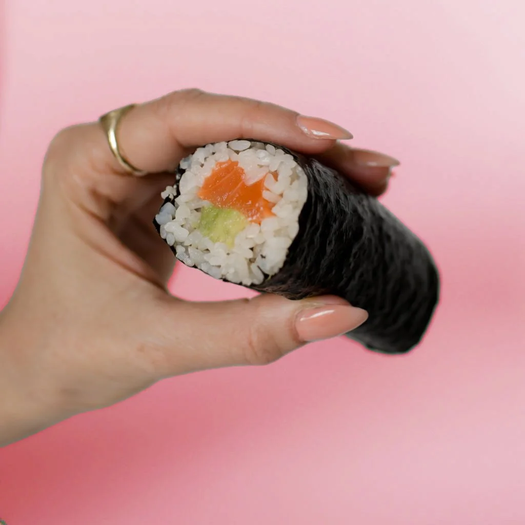 Close-up of a hand holding a sushi roll with visible ingredients of salmon and cucumber inside, set against a pink background.