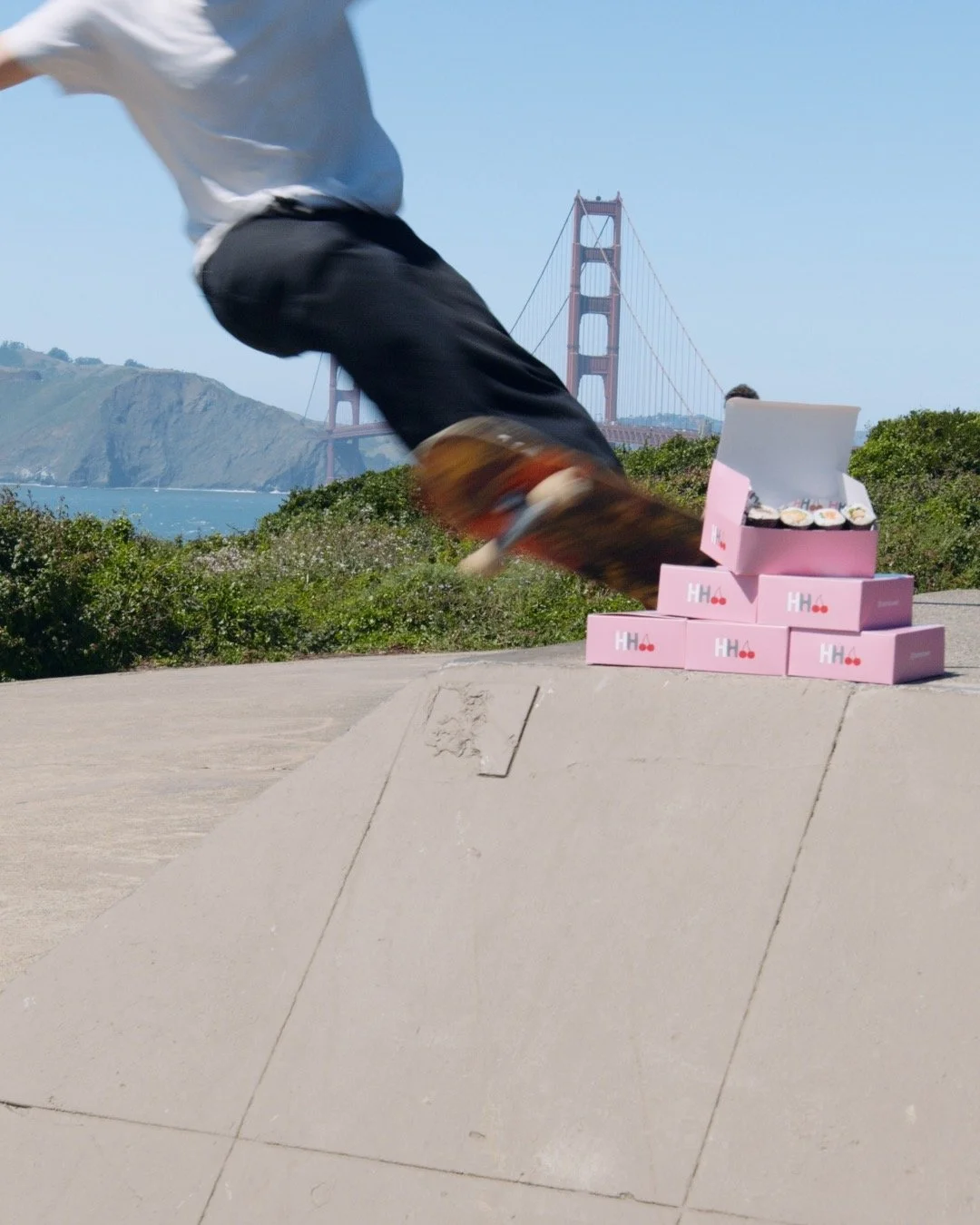 A skateboarder is in mid-air performing a trick at a skate park, with a stack of pink boxes labeled 'H H' and a box of sushi in front of him. The Golden Gate Bridge and hills are visible in the background on a sunny day.